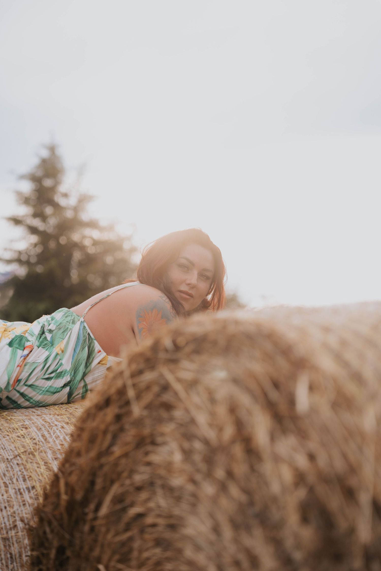 A woman in a floral dress rests on a large hay bale in a rural field, gazing at the camera with a serene expression.