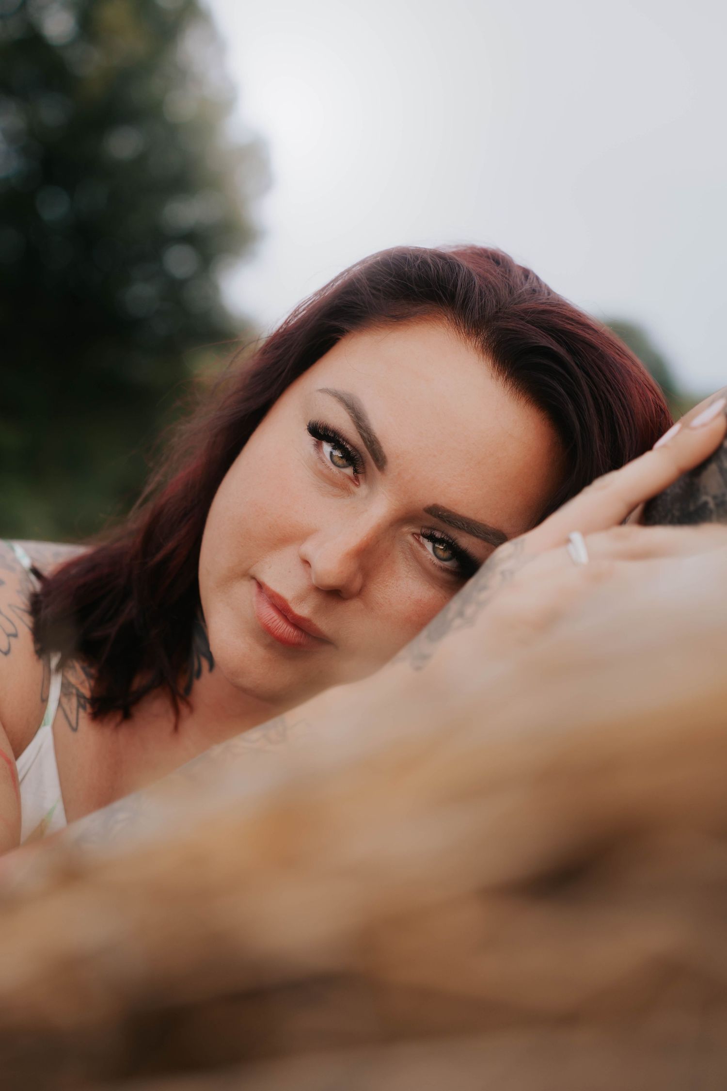 A woman with red hair and tattoos rests her head on a rock, gazing softly at the camera outdoors.