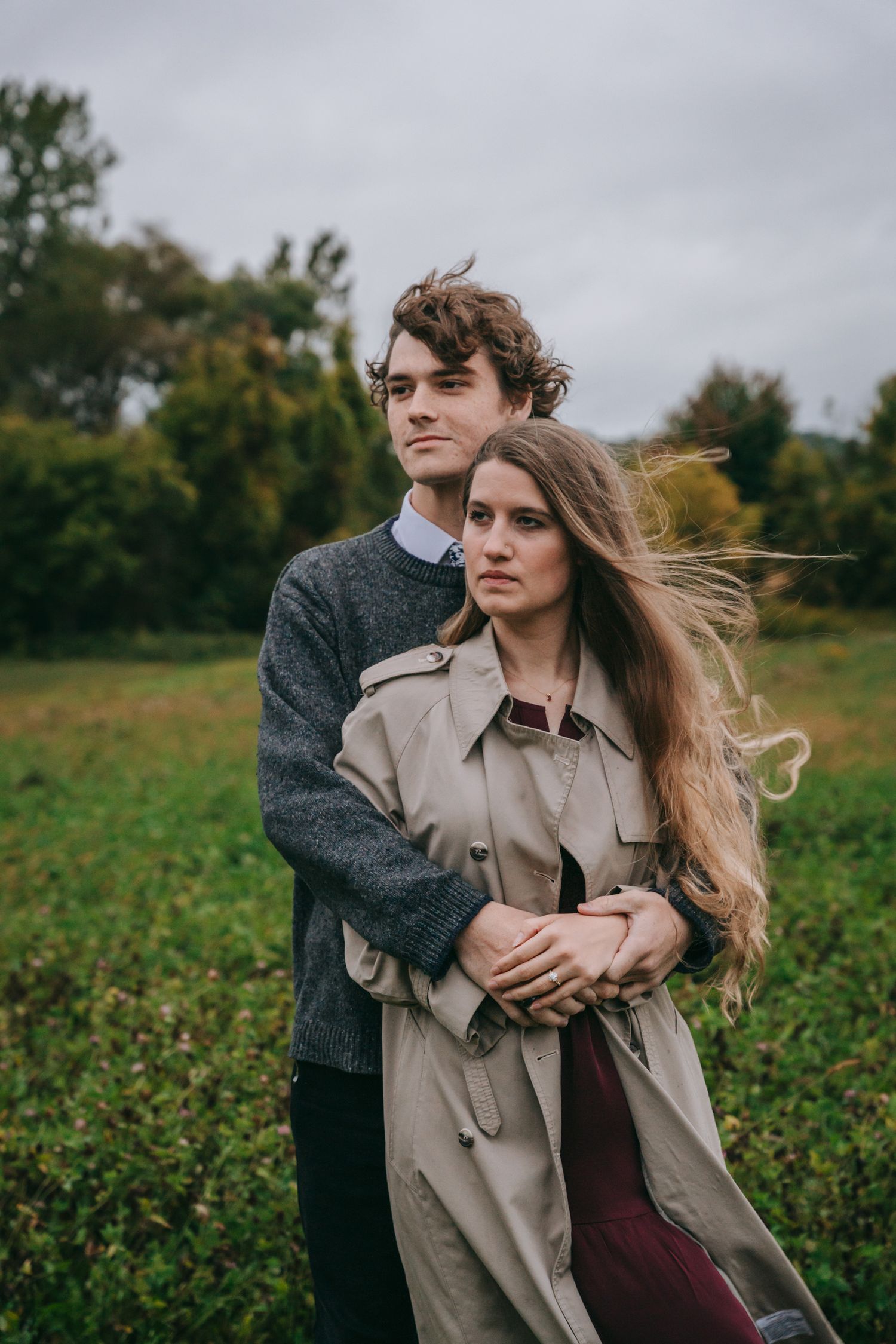 Engaged couple standing together in Finger Lakes wine country, holding each other in an open green field with autumn foliage in background