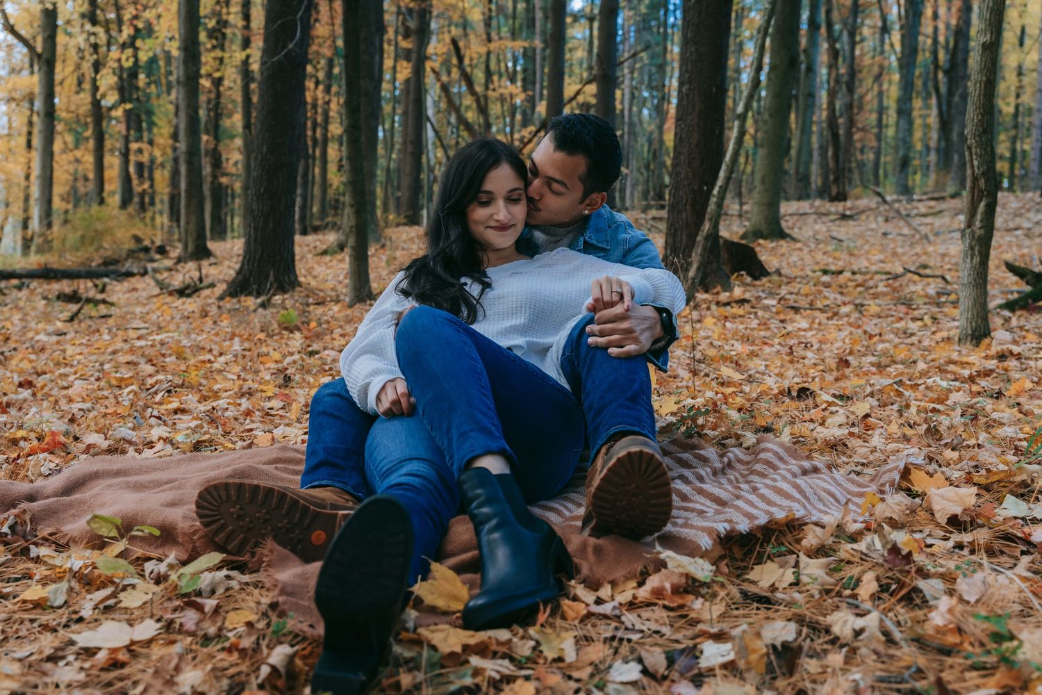 Couple cuddling on a blanket in autumn woods at Letchworth State Park, surrounded by fallen leaves during a cozy fall engagement session