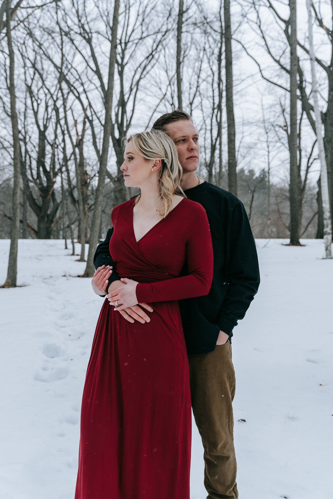 Engaged couple standing together in snowy woods, woman in flowing red dress during winter engagement session in Rochester NY
