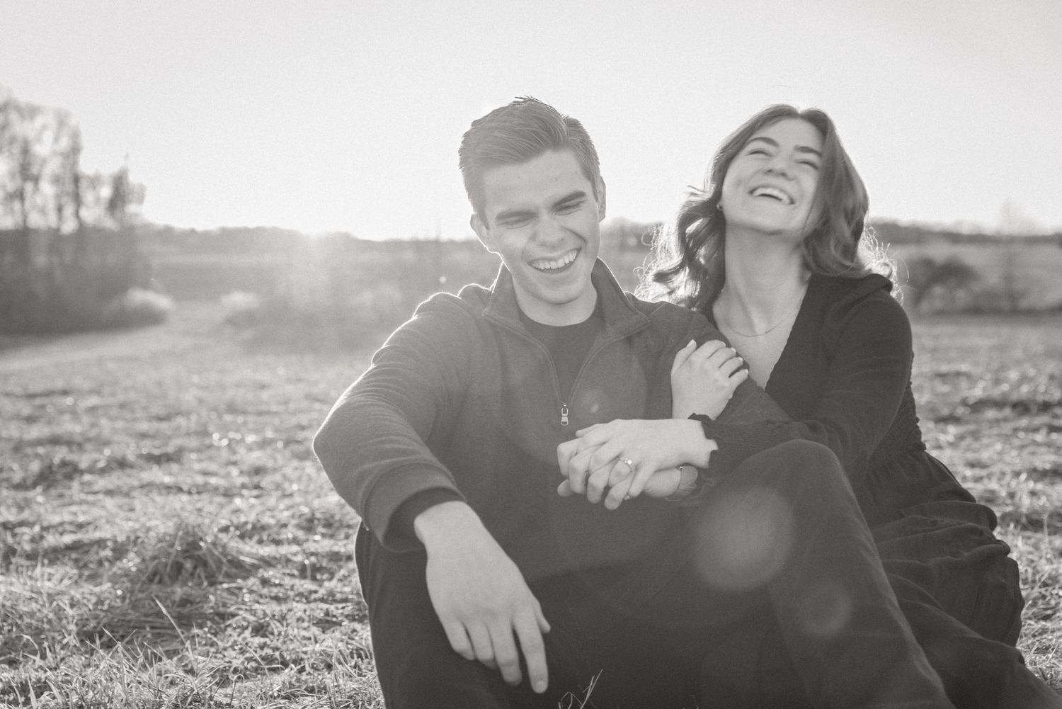 Black and white engagement photo of a laughing couple sitting together in an open Finger Lakes field