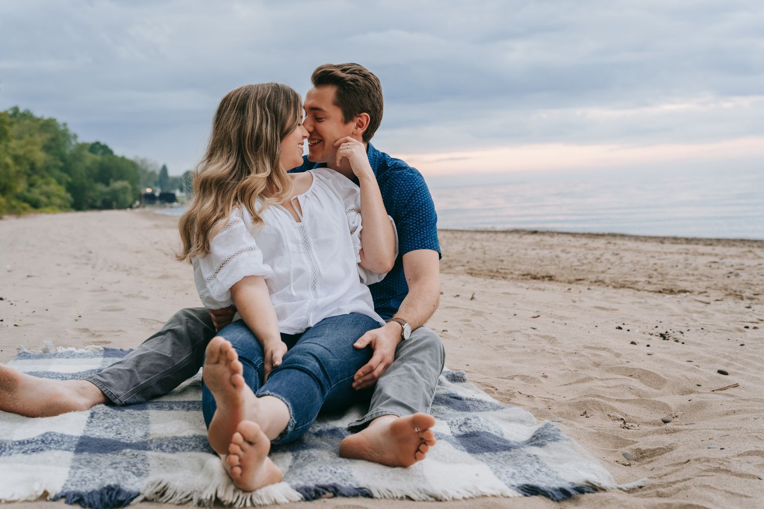 Engaged couple sitting barefoot on a blanket on a sandy Lake Ontario beach near Rochester, NY, laughing and leaning in together at sunset