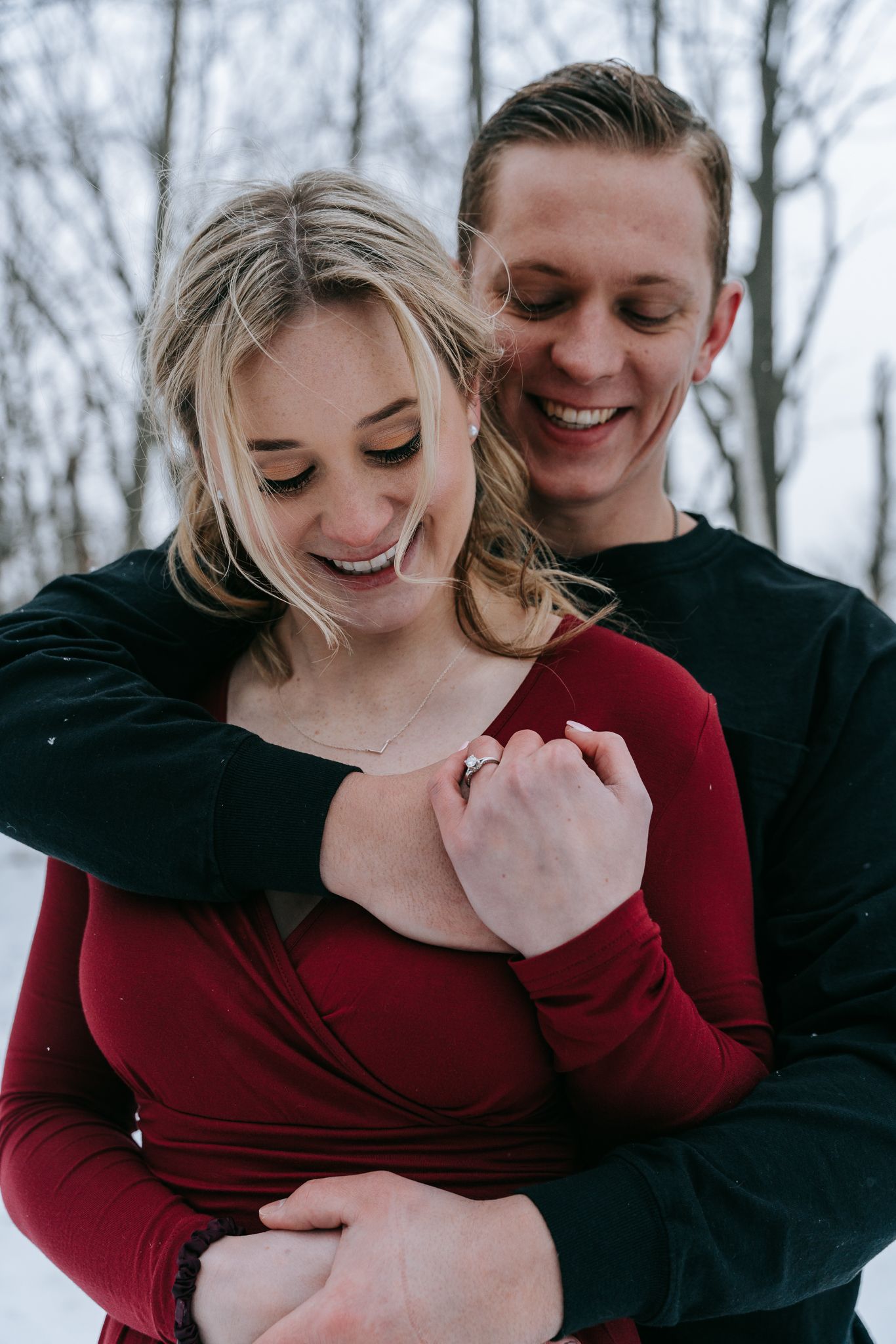 Engaged couple embracing in snowy winter, engagement ring visible as they smile together during candid Rochester NY engagement session