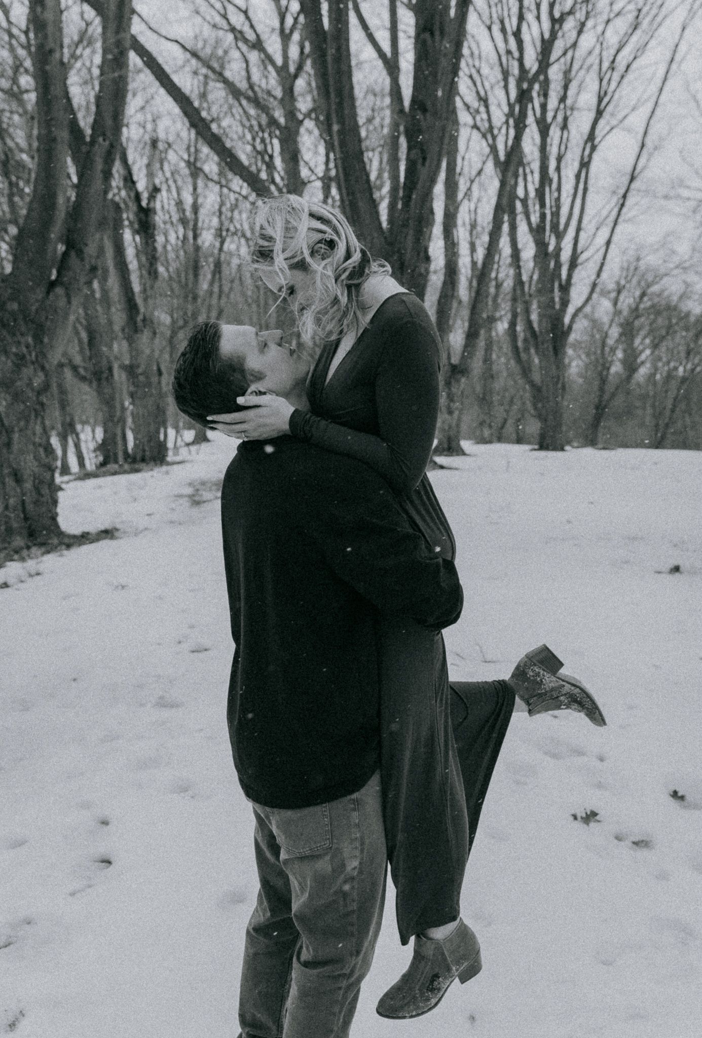 Engaged couple embracing in snowy forest, black and white winter engagement photo with intimate documentary style, Rochester NY park