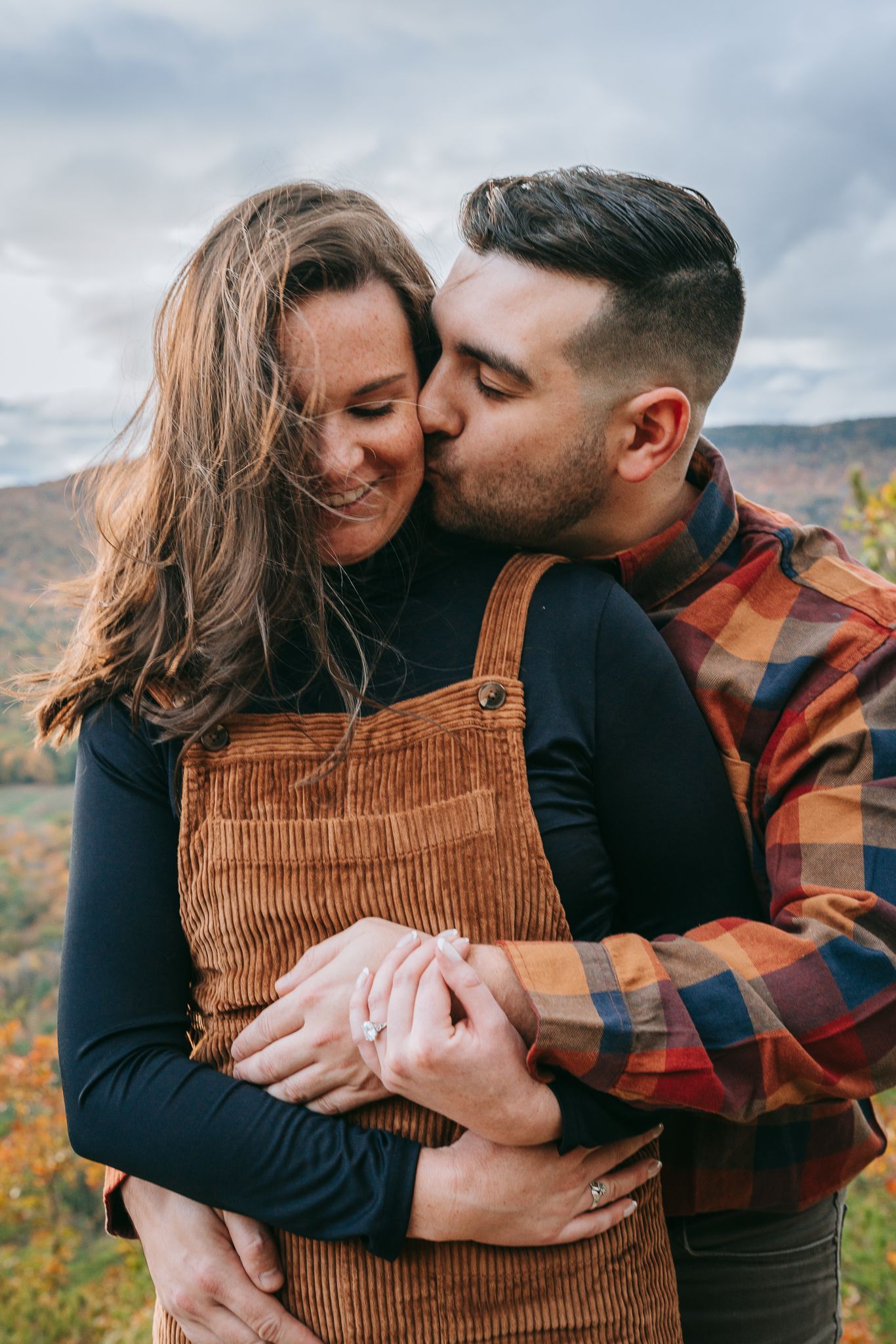 Closeup engagement portrait of couple embracing on a mountaintop in the White Mountains of New Hampshire, with fall foliage in the distance