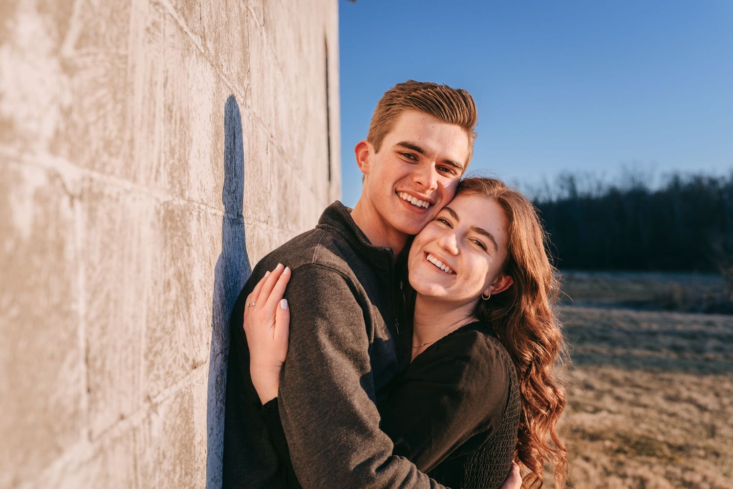 Smiling engaged couple leaning against a barn in the Finger Lakes, captured in warm evening light