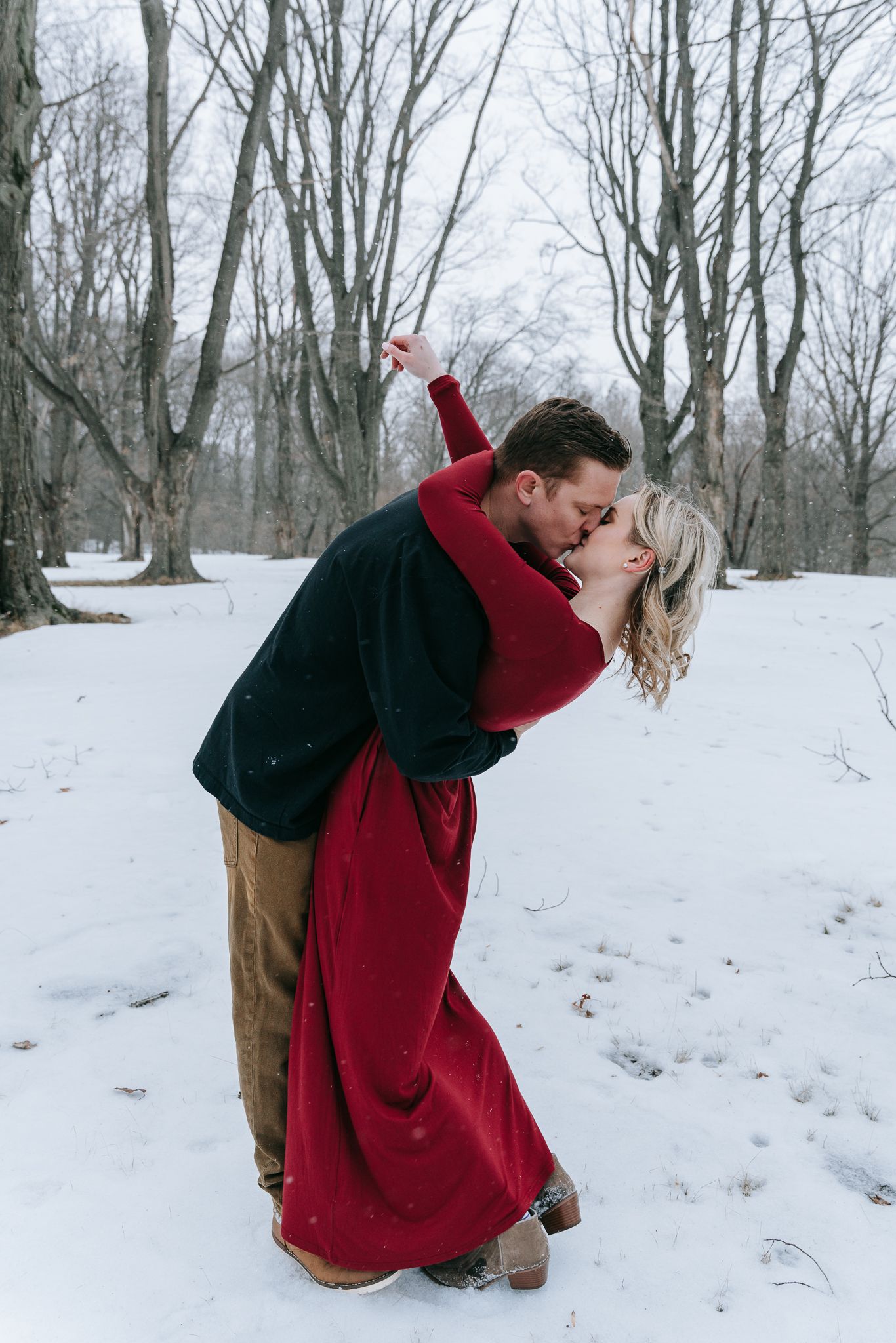 Couple kissing in snowy Rochester NY, woman in red dress and man in black coat, romantic engagement photography in Upstate NY, snowy session