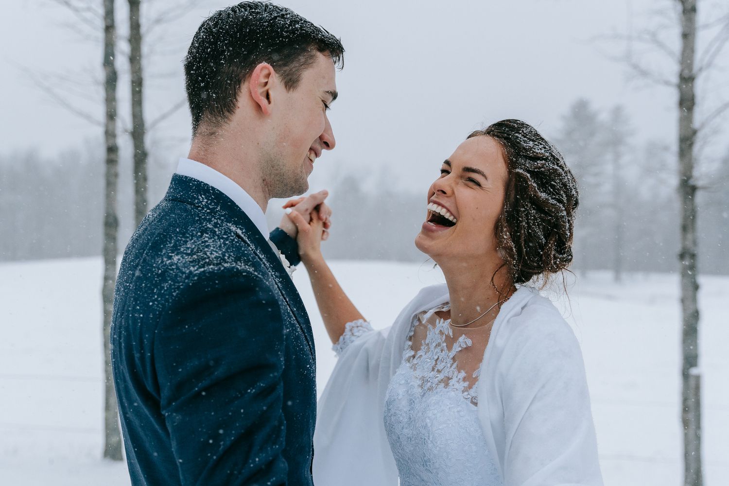 Bride and groom laughing together as snow falls at winter wedding in the Green Mountains VT, candid documentary wedding photography style