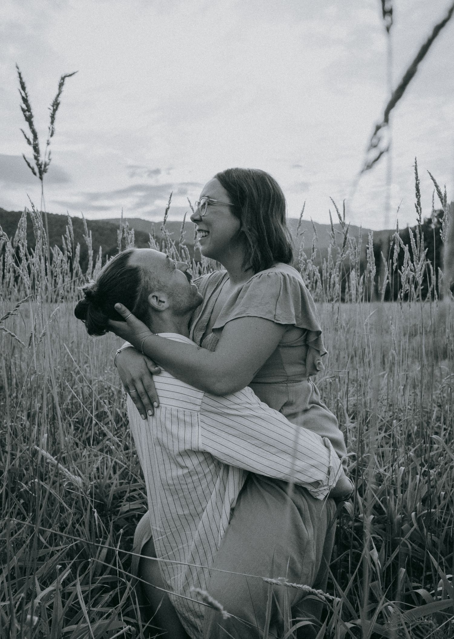 Engaged couple embracing and laughing in tall grass during black and white engagement session in Keene Valley, Adirondack High Peaks region