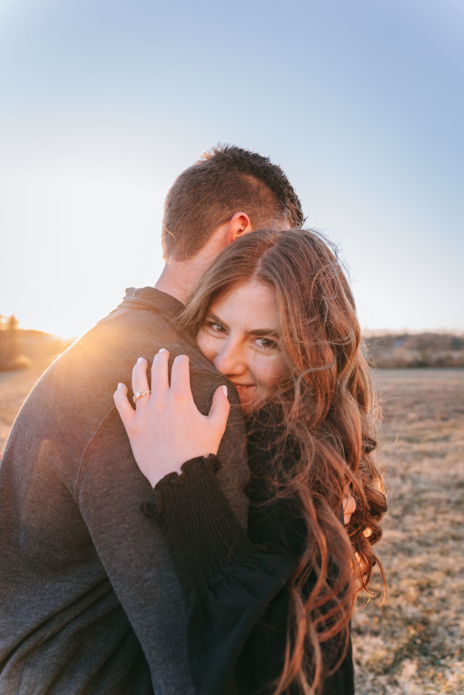 Engaged couple embracing at sunset in a Finger Lakes field, golden light highlighting her engagement ring and intimate moment
