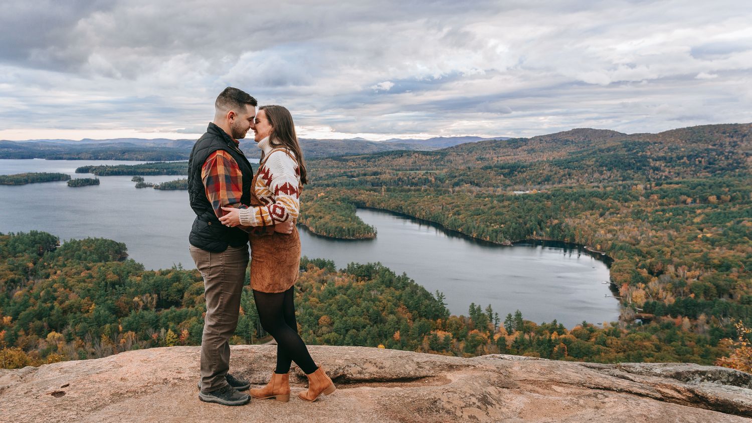 Wide scenic engagement portrait of a couple standing on a rocky overlook above Squam Lake, surrounded by autumn colors in the White Mountain