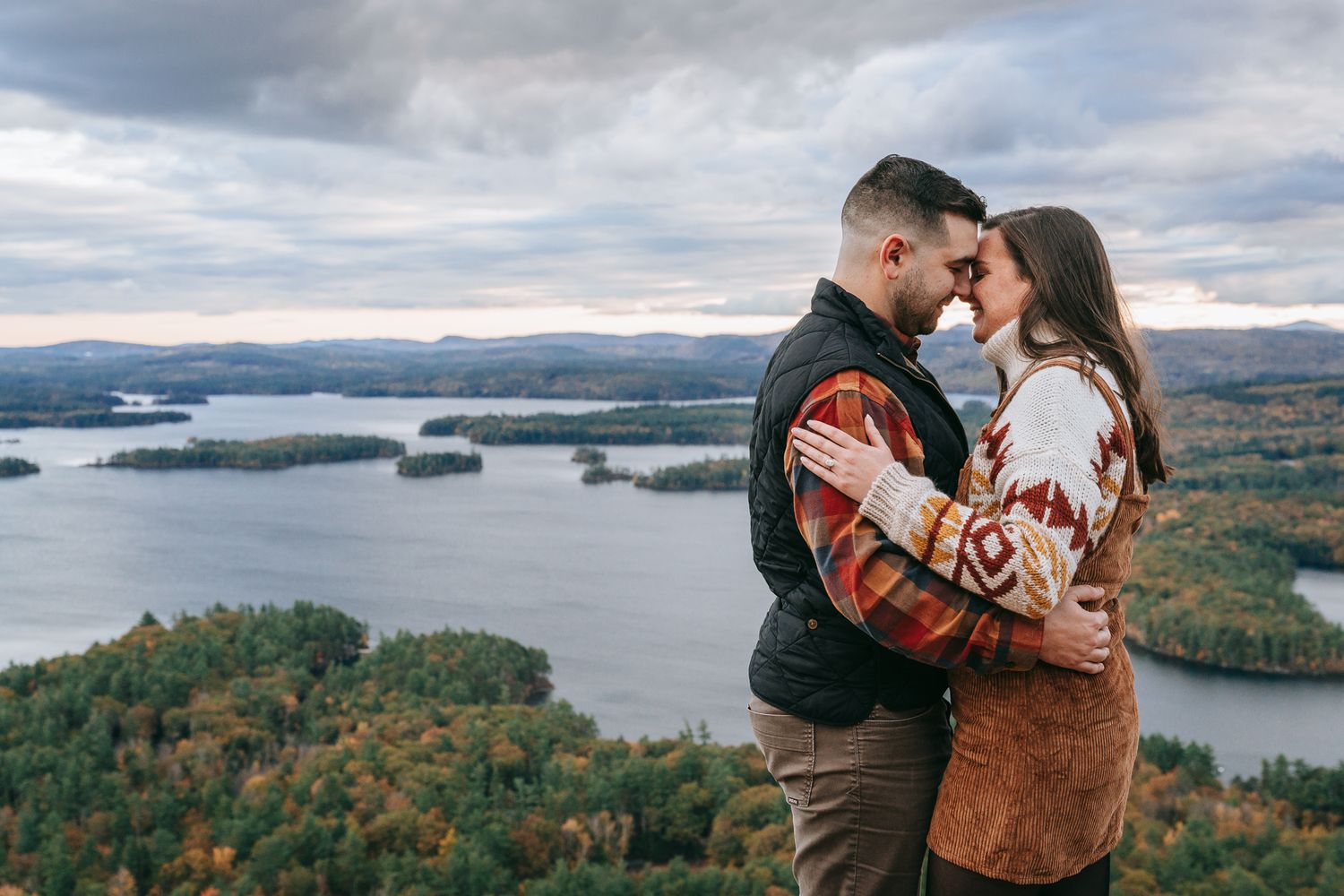 Couple embracing with scenic White Mountains NH mountain views behind them during adventure engagement session, candid NH photography