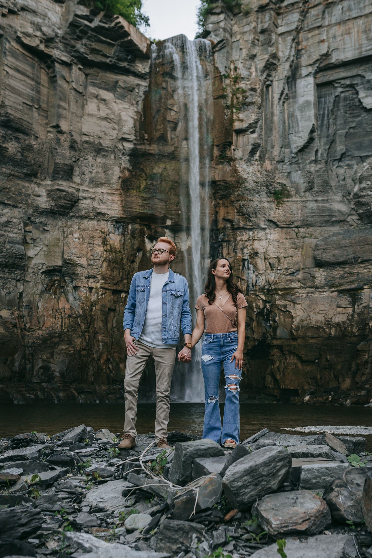 Engaged couple holding hands in front of Taughannock Falls waterfall at Taughannock Falls State Park in the Finger Lakes near Ithaca, NY