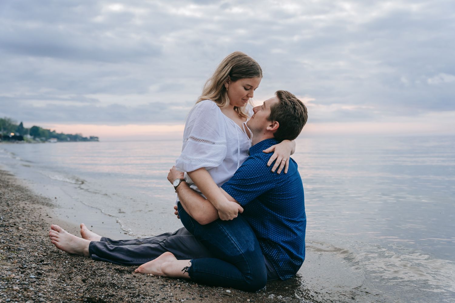 Romantic engagement photo of couple sitting at water’s edge on a Lake Ontario beach near Rochester NY, holding each other during golden hour