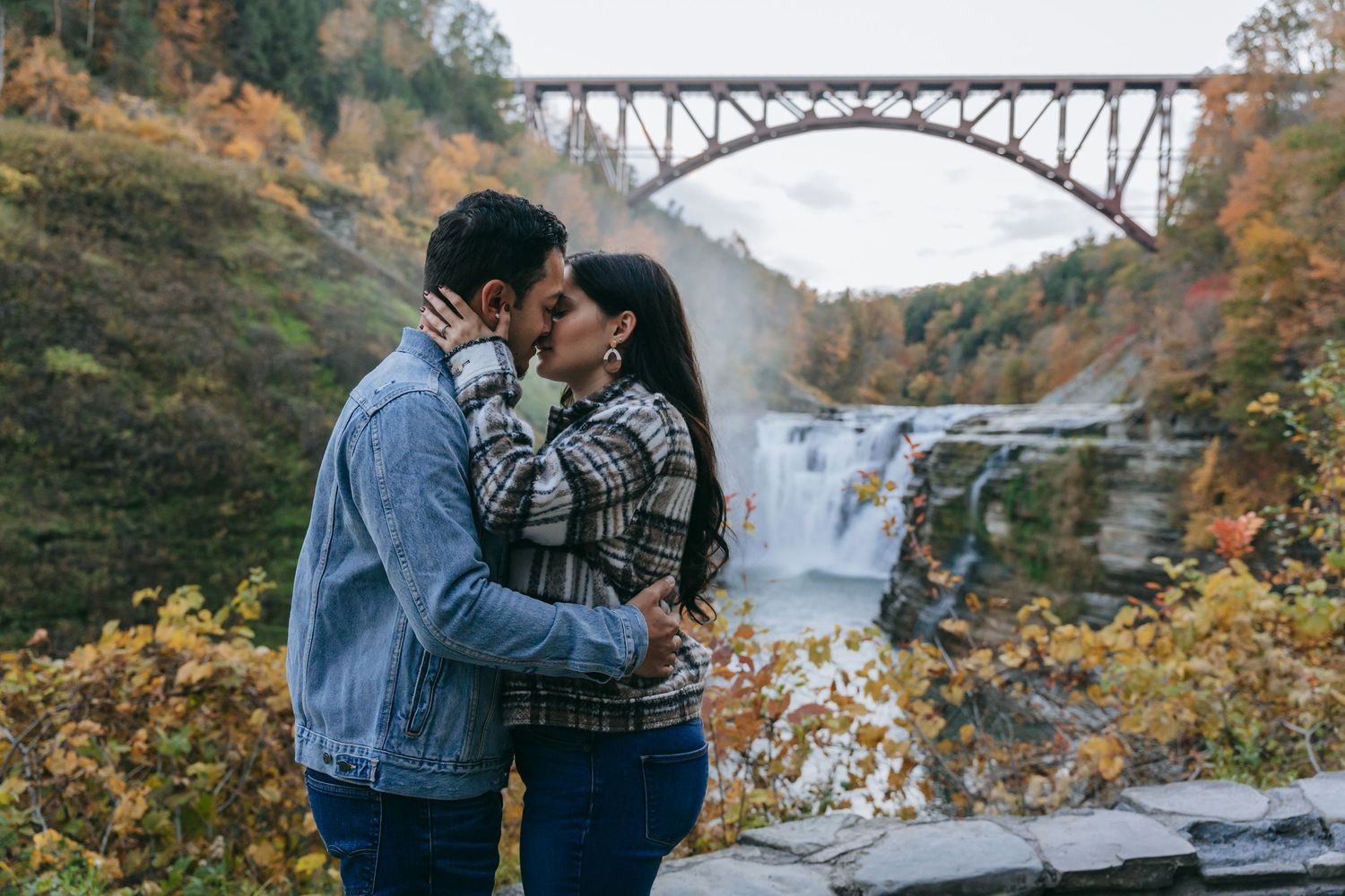 Engaged couple kissing near a waterfall at Letchworth State Park in New York, framed by fall foliage and the iconic Upper Falls bridge