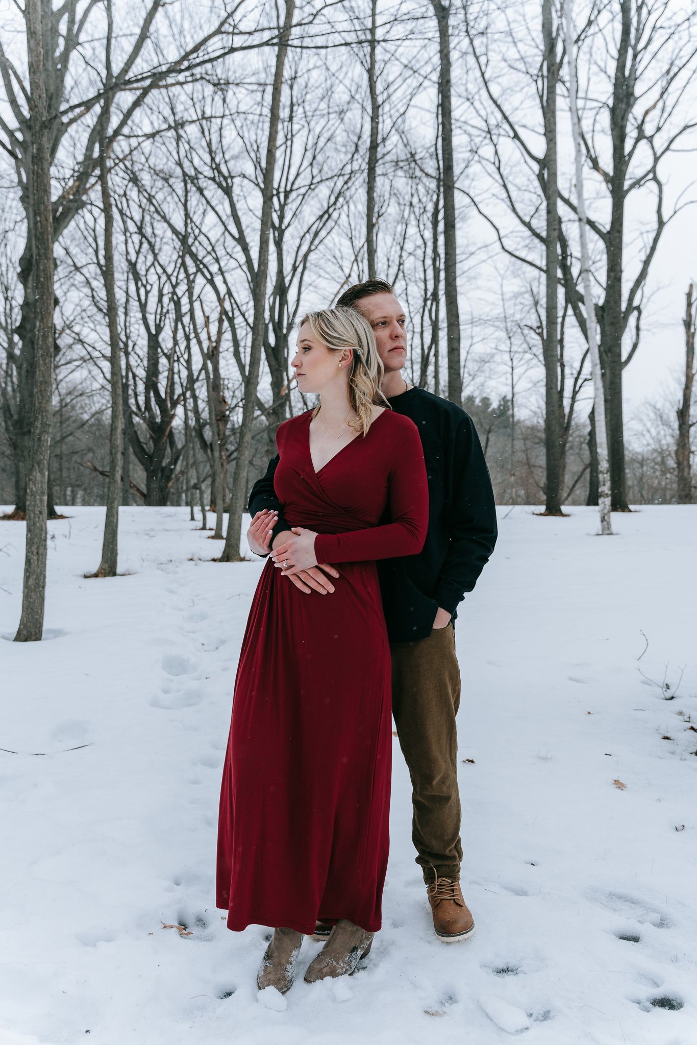 Engaged couple standing together in snowy woods, woman in flowing red dress during intimate romantic winter engagement session in Upstate NY