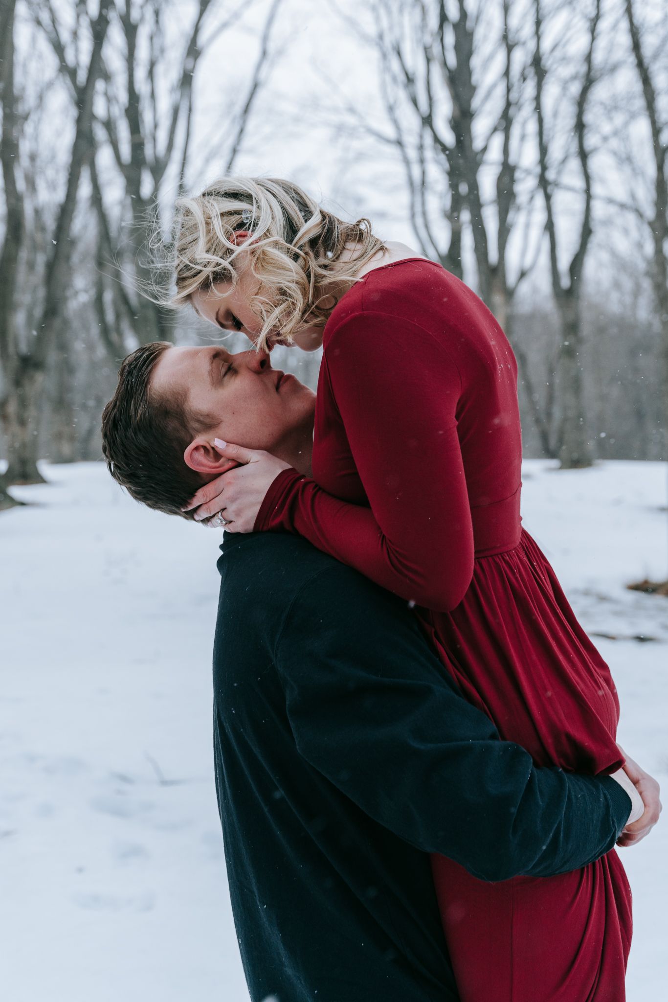 Engaged couple intimate embrace in falling snow, woman in red dress lifted by man during romantic winter engagement session in Rochester NY