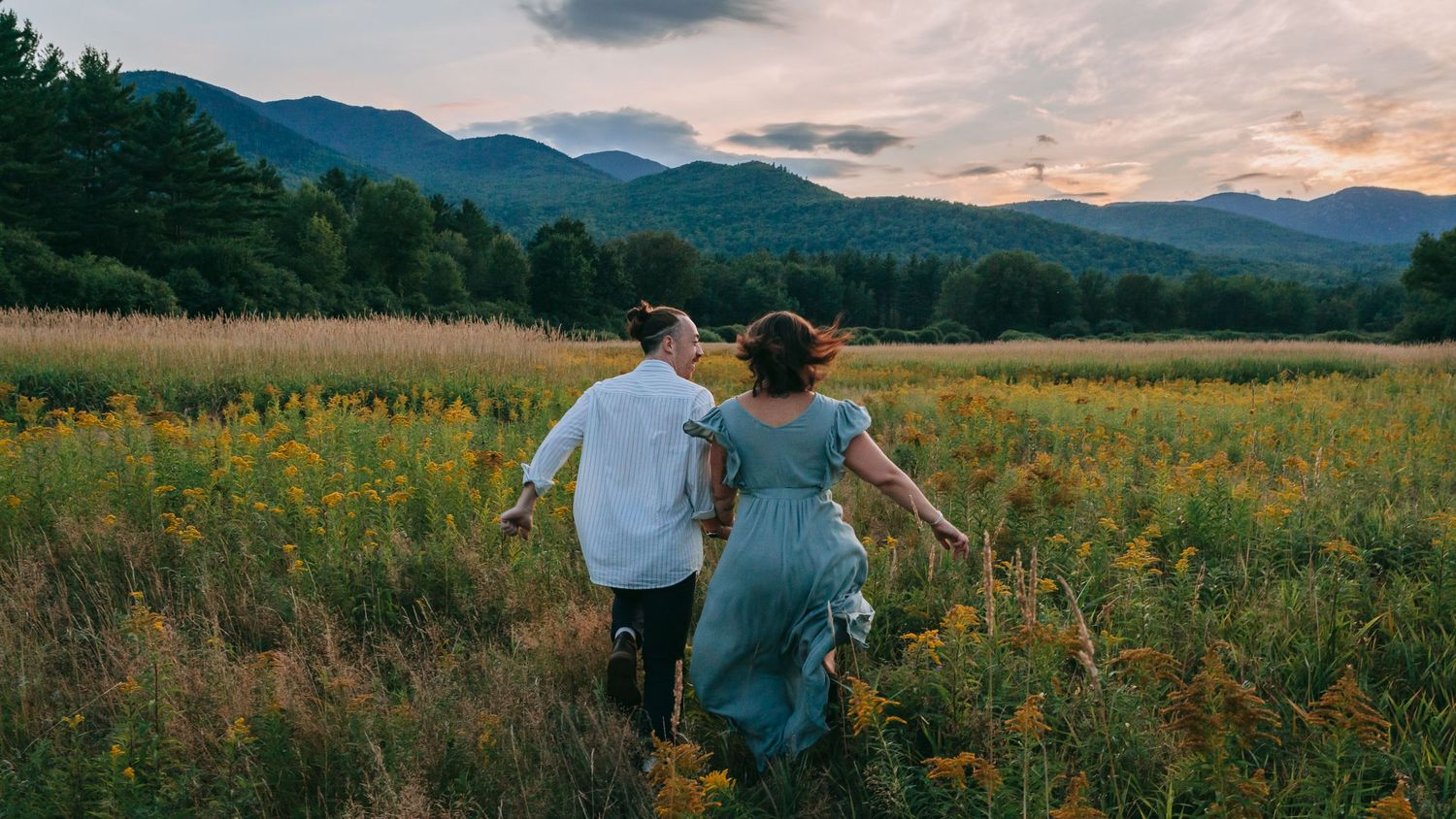 Couple running through goldenrod wildflower field with Adirondack High Peaks in background during Keene Valley engagement session at sunset