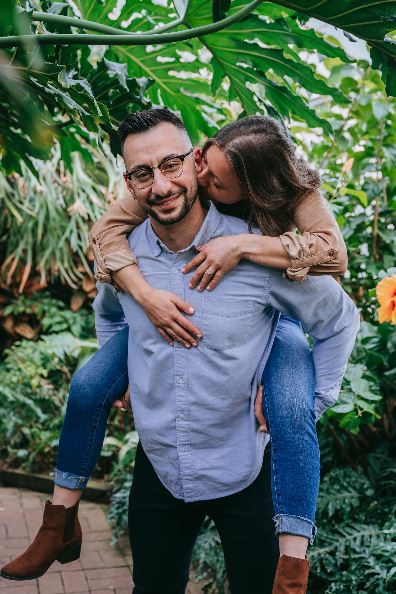 Engaged couple sharing playful piggyback moment inside Lamberton Highland Park Conservatory in Rochester NY, candid engagement photography