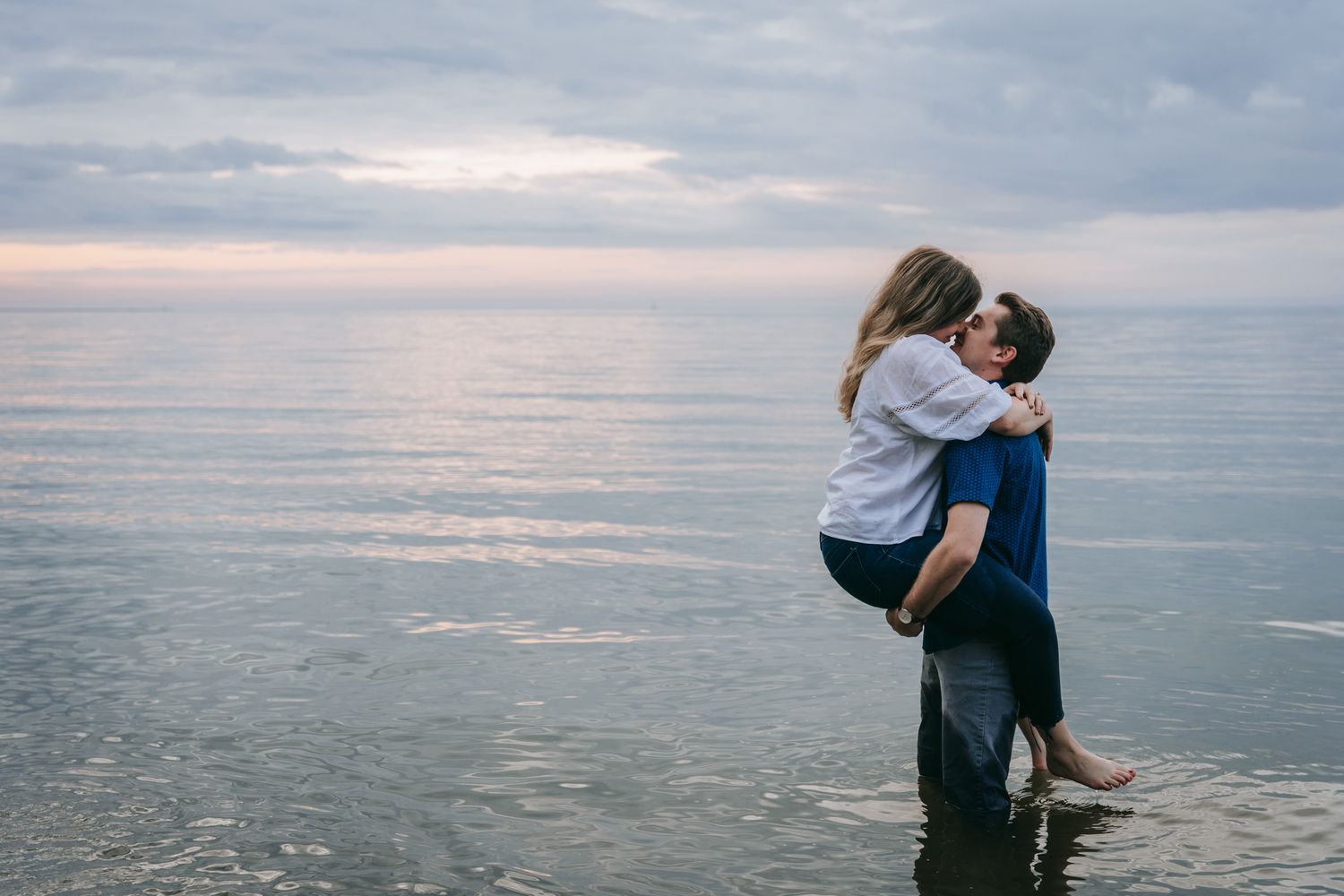 Couple embracing and kissing while standing in shallow water along Lake Ontario near Rochester, New York, with calm waves and pastel evening