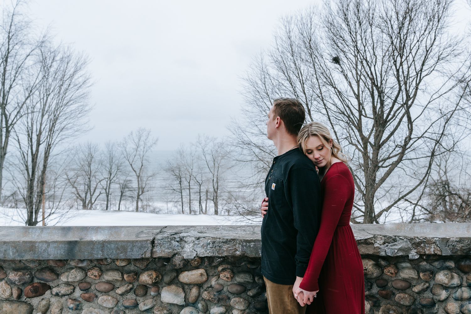 Engaged couple holding hands by stone wall in winter, woman in red dress leaning into fiance during Rochester NY engagement session