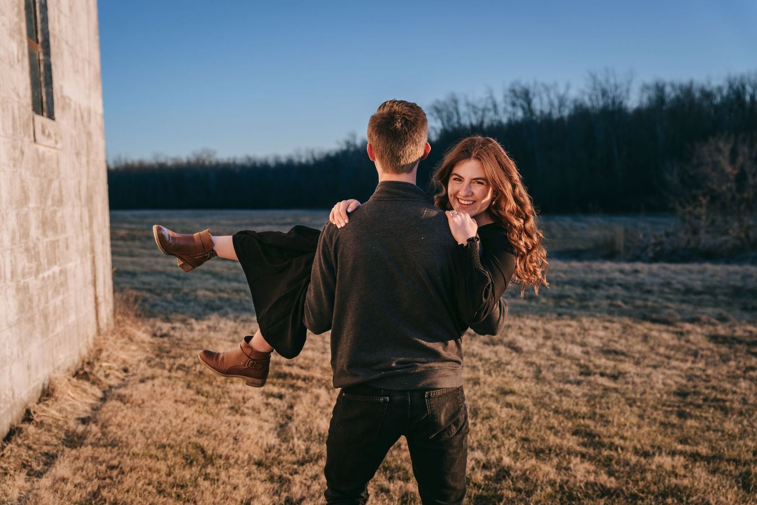 Joyful engagement photo in the Finger Lakes with groom lifting bride near a rustic barn during golden hour