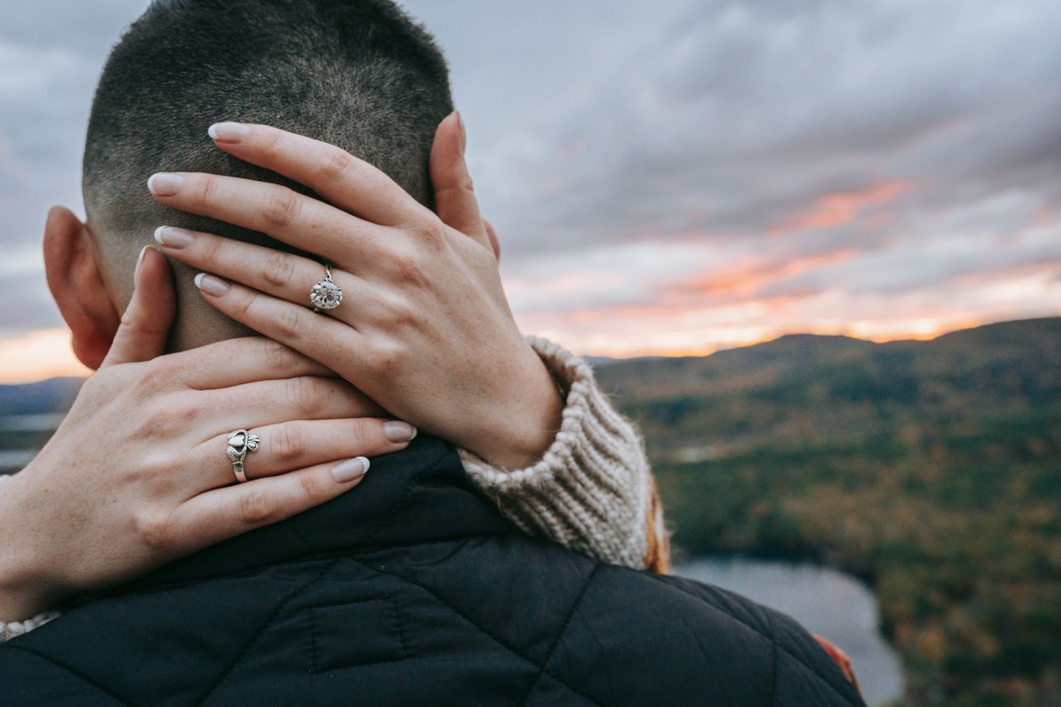 Detail engagement photo of hands wrapped around a man’s neck, featuring an engagement ring at sunset above Squam Lake in the White Mount