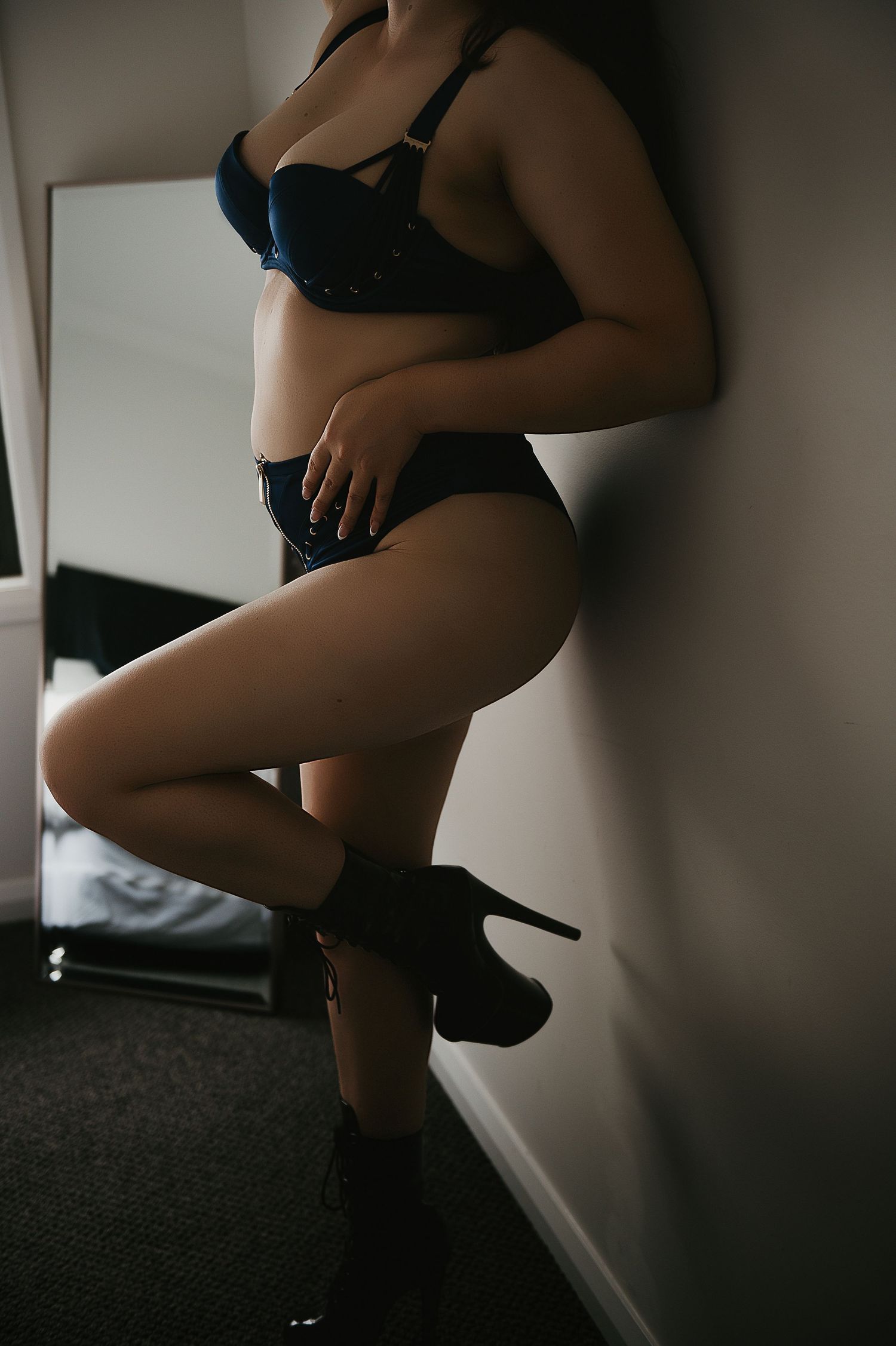 Woman in dark blue lingerie and high heels posing against a white wall in a dimly lit room for boudoir photographer SeduXion Studio in Perth