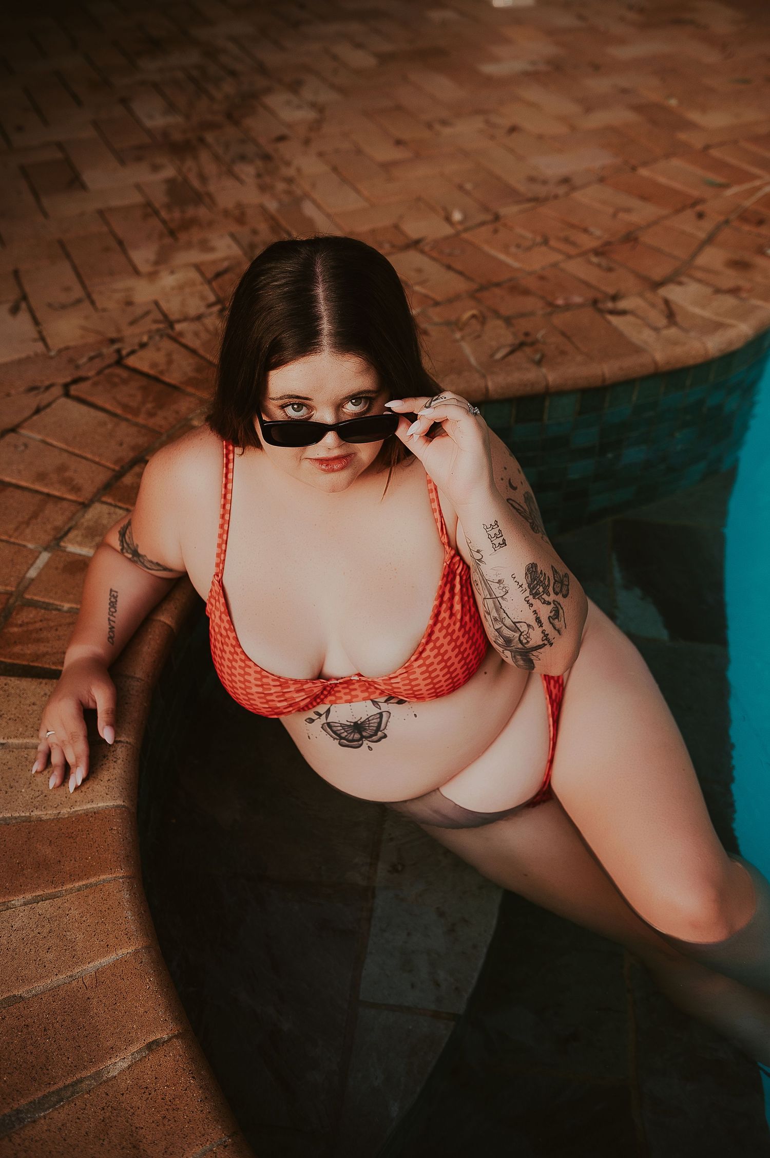Woman in red bikini with tattoos and sunglasses posing by a pool on brick pavement. Boudoir Photographer Perth WA. SeduXion Studio Mandurah