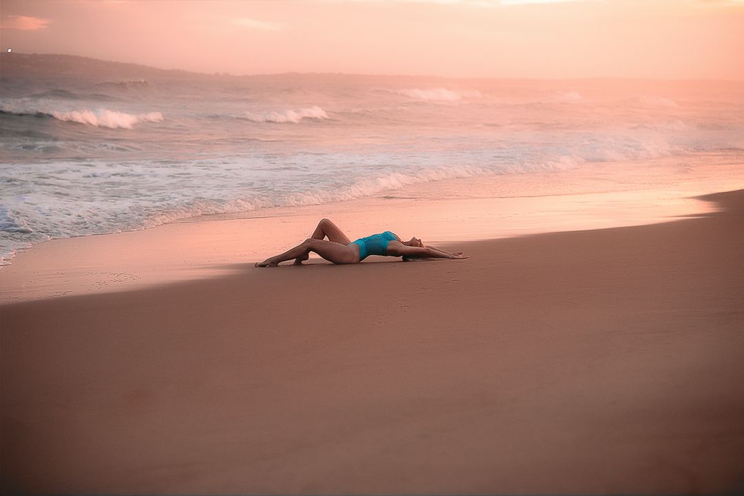 A woman lies on a sandy beach at sunset creating a silhouette against the pink and orange sky for boudoir photoshoot in Mandurah WA