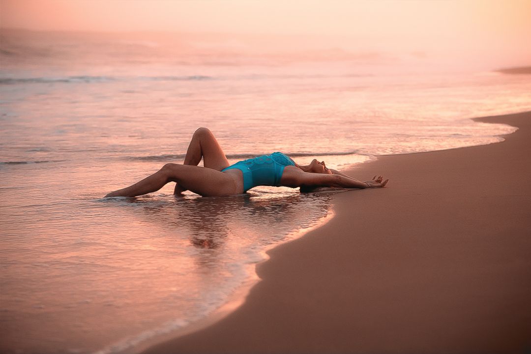 A woman lies in shallow ocean water during sunset for boudoir photoshoot, creating a serene silhouette against the pink and orange sky.