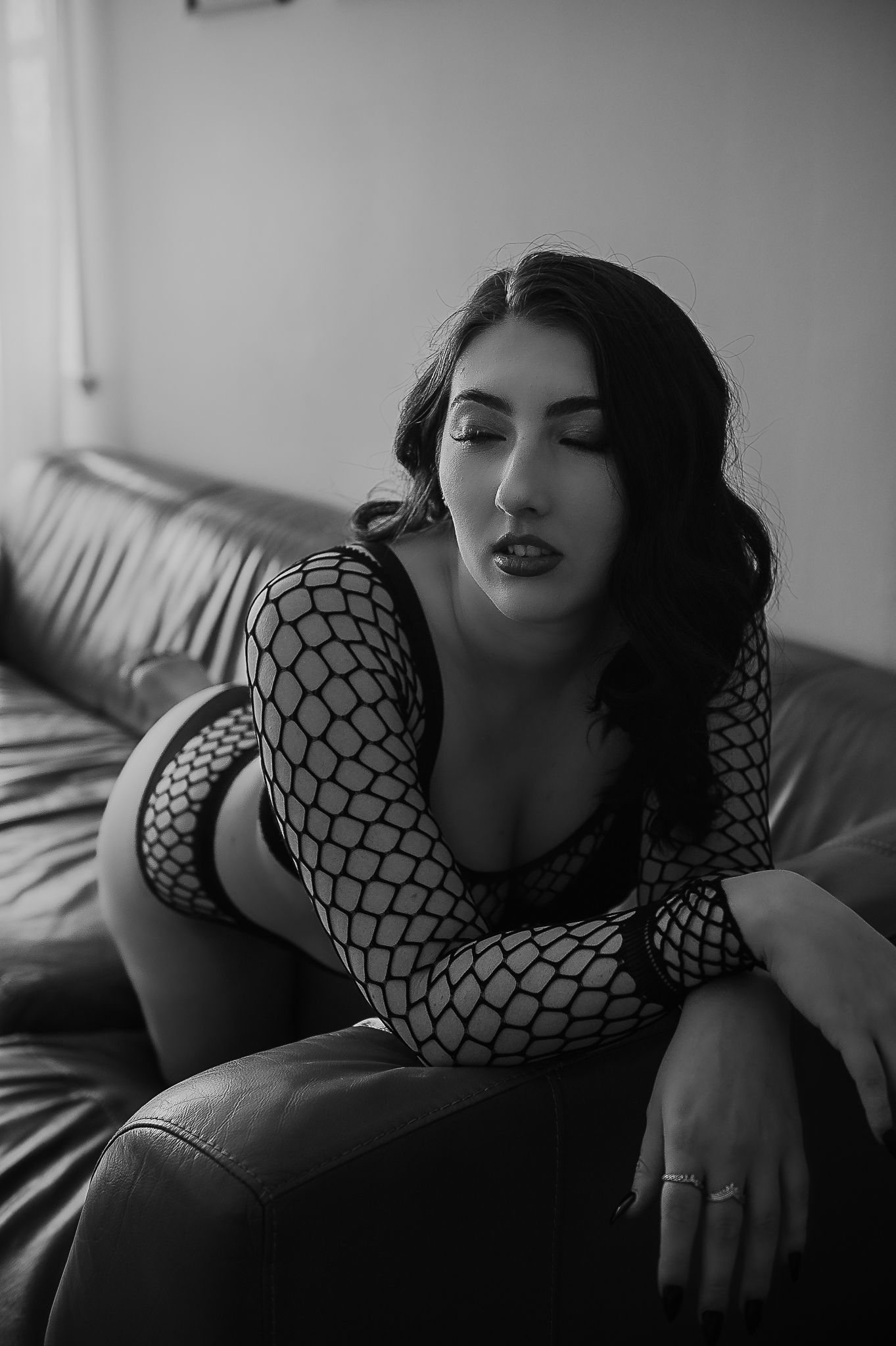 A black and white boudoir photo showing a woman in fishnet sleeves reclining on a couch at SeduXion Studio in Mandurah Western Australia