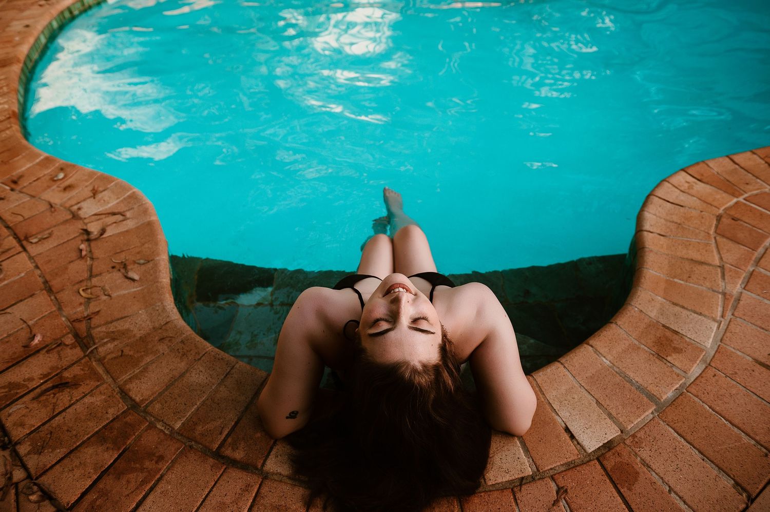 A woman poses in a black swimsuit on the edge of the blue pool for boudoir photographer SeduXion Studio in Mandurah WA