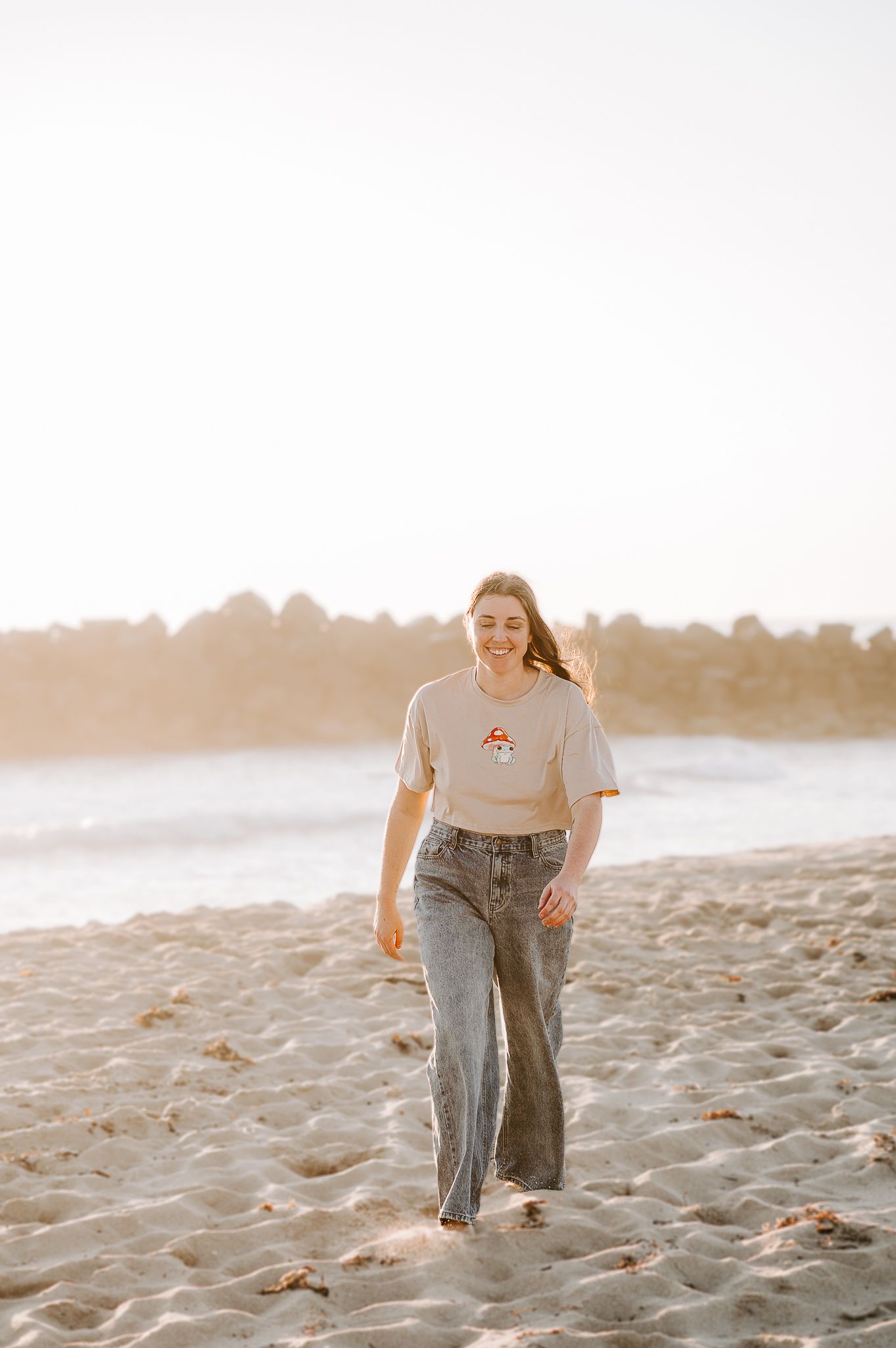 Woman walking on a sandy beach at sunset wearing a casual t-shirt and wide-leg jeans. SeduXion Studio in Mandurah Western Australia