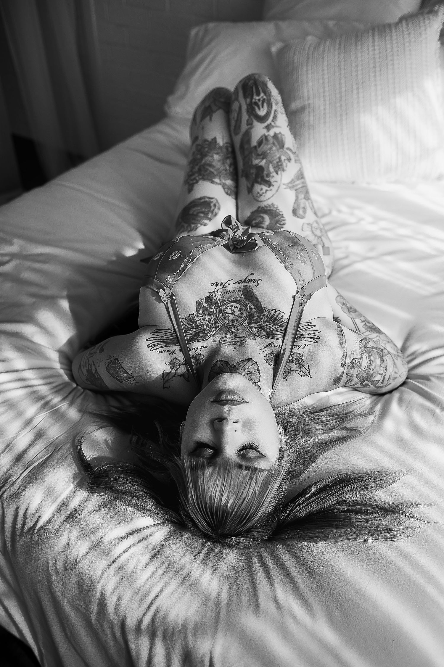 Black and white photo of a tattooed woman lying on a white bed posing for boudoir photographer SeduXion Studio in Mandurah Western Australia