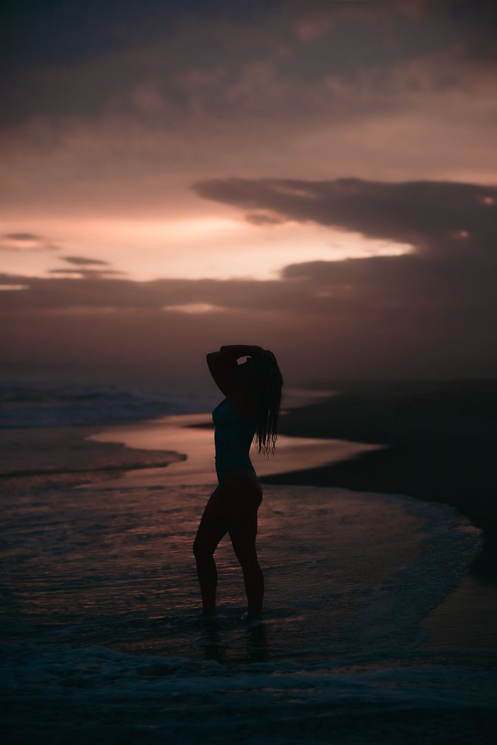 Silhouette of woman standing on beach at sunset against a dramatic cloudy sky with dark moody atmosphere for boudoir photos in Mandurah WA