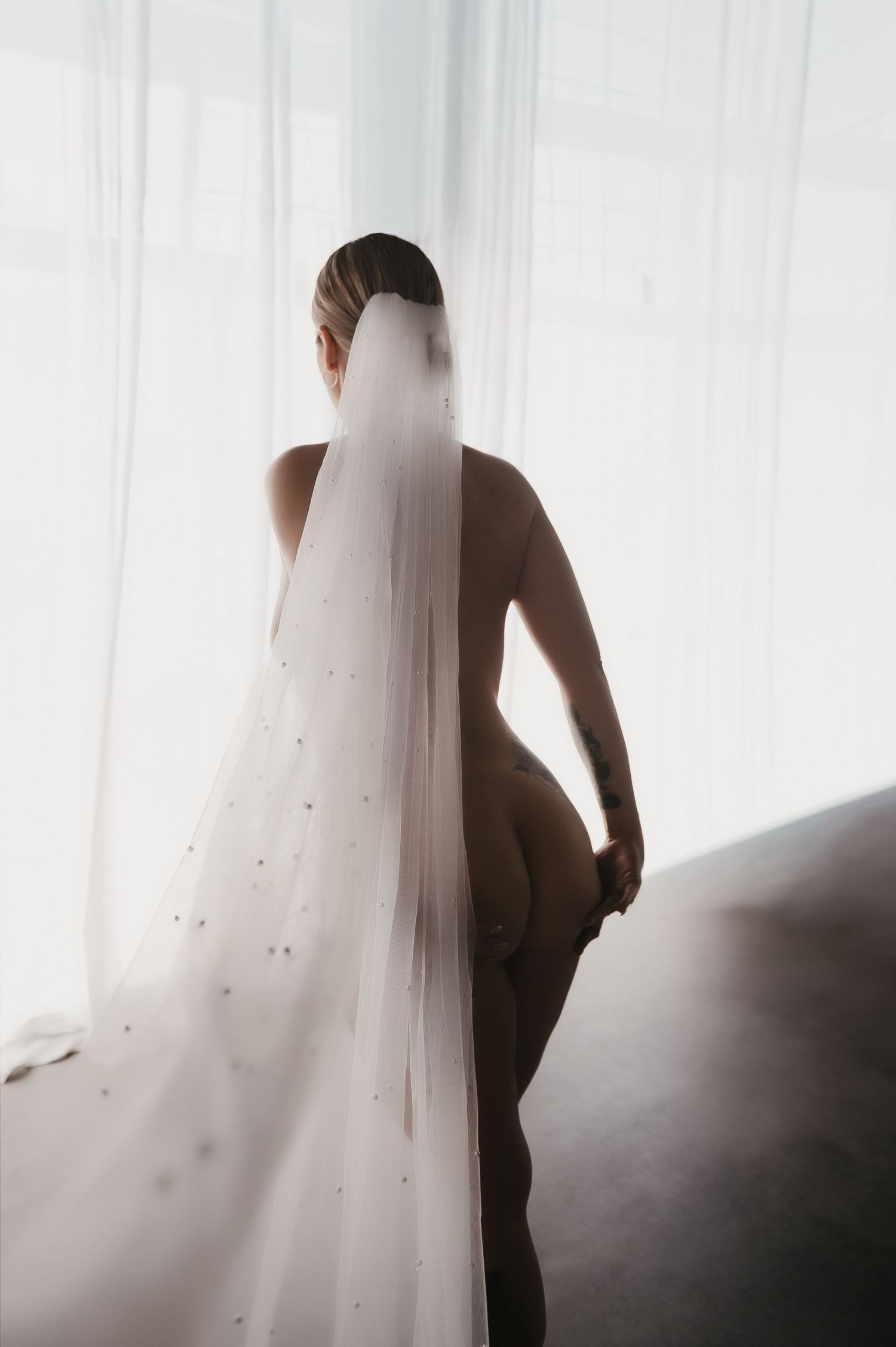 A bride wearing only a pearl-embellished wedding veil stands backlit by sheer curtains in an artistic boudoir photoshoot at SeduXion Studio