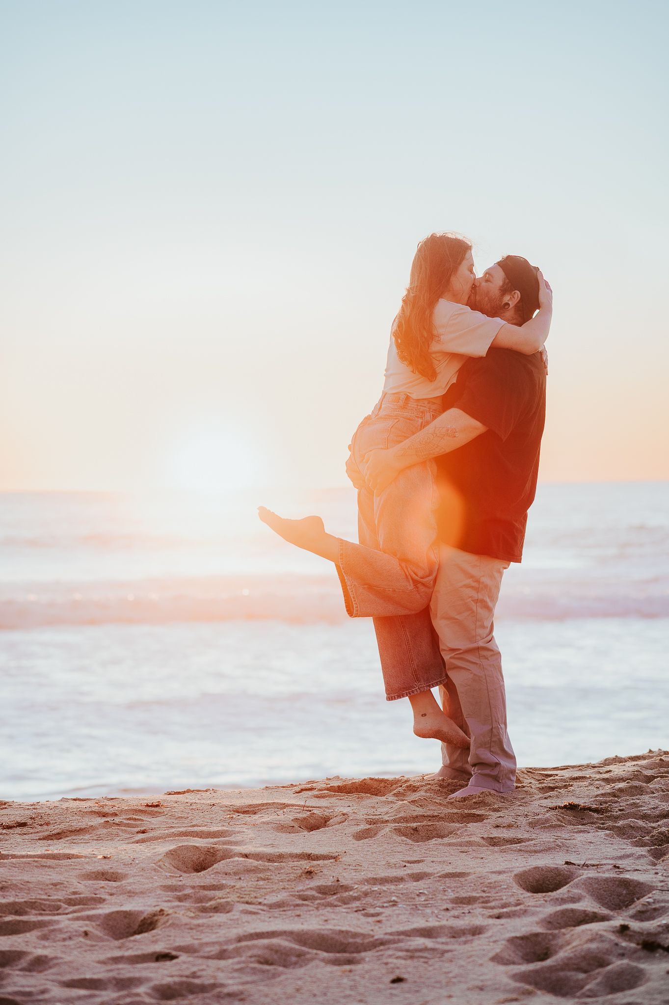 A couple embraces on a sandy beach at sunset with warm golden light creating a romantic silhouette. SeduXion Studio Mandurah WA
