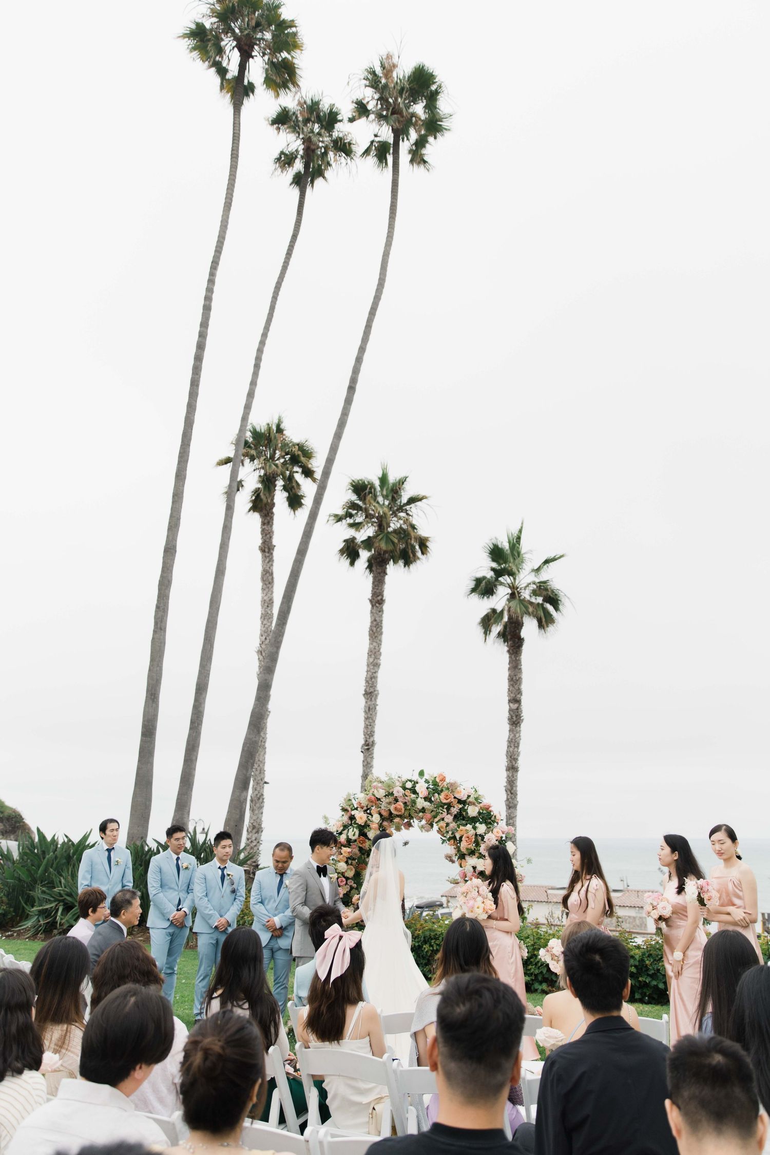 Wedding ceremony scene with tall palm trees and floral arch overlooking the ocean.