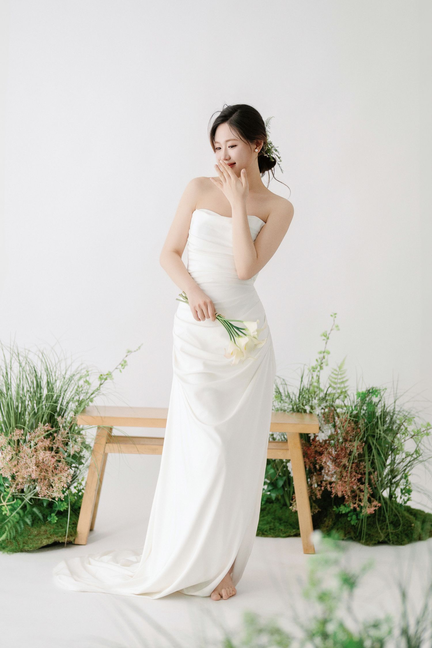 Artistic bridal session with wooden bench and potted greenery against white studio backdrop.