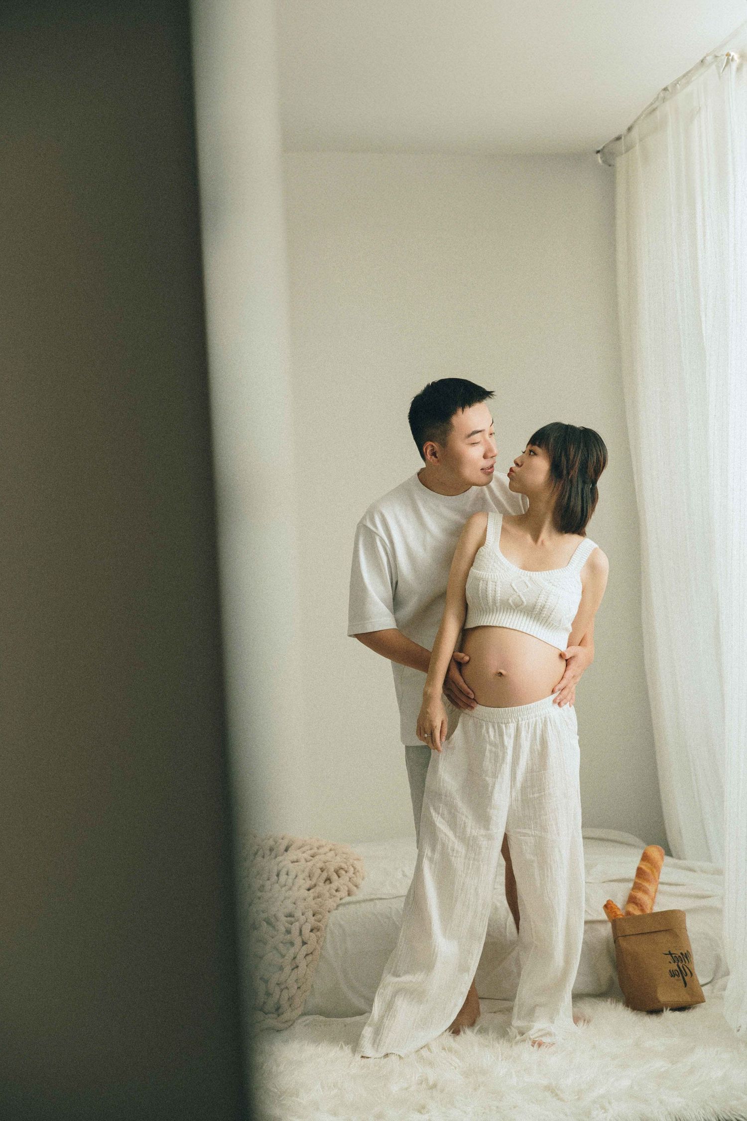 Couple in white clothing posing together near window with soft natural lighting.