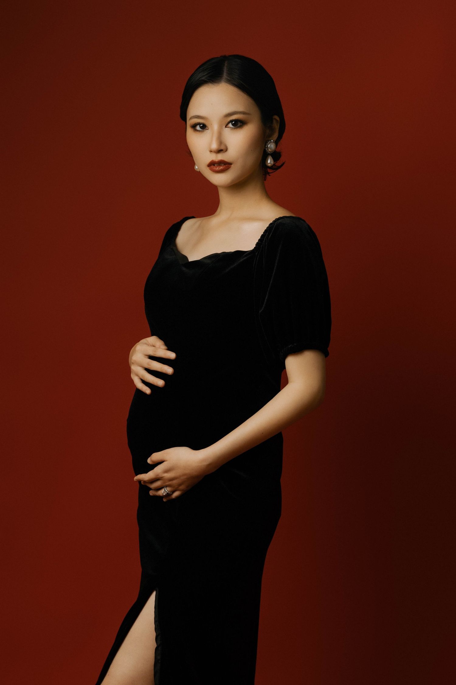 Dramatic maternity portrait against burgundy backdrop wearing elegant black dress.