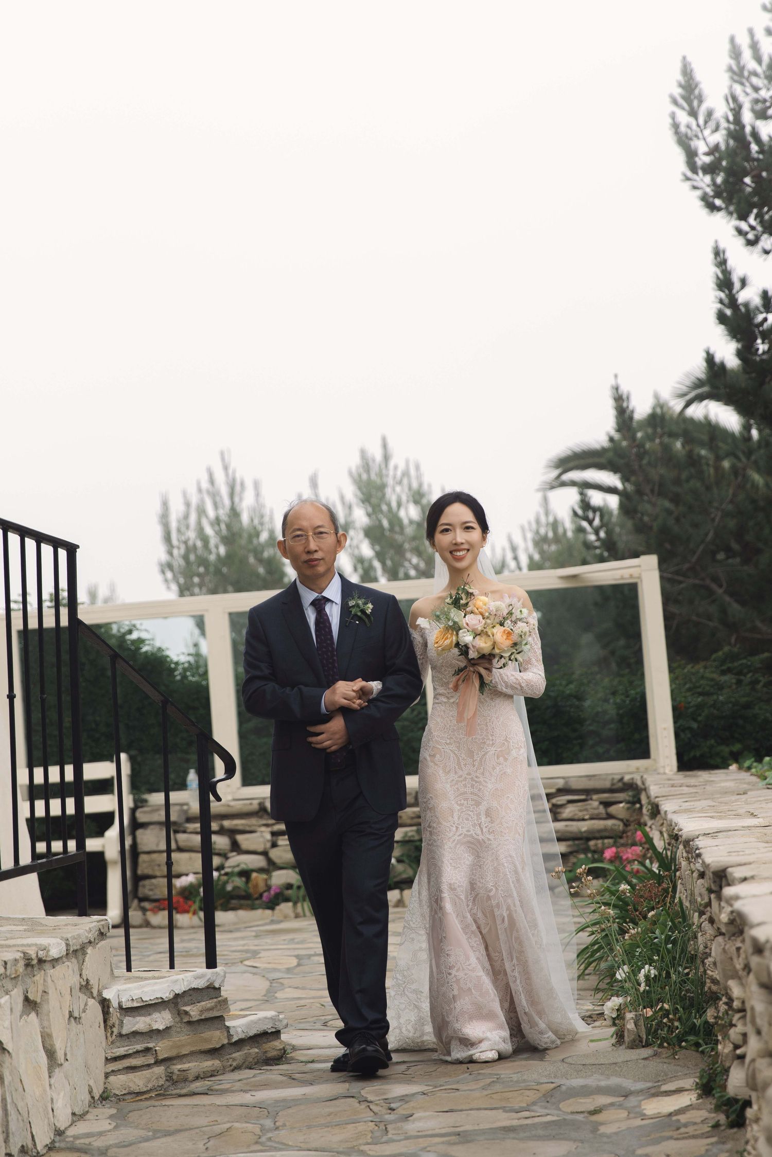 Wedding party members walk and pose on a stone patio with greenery in the background.