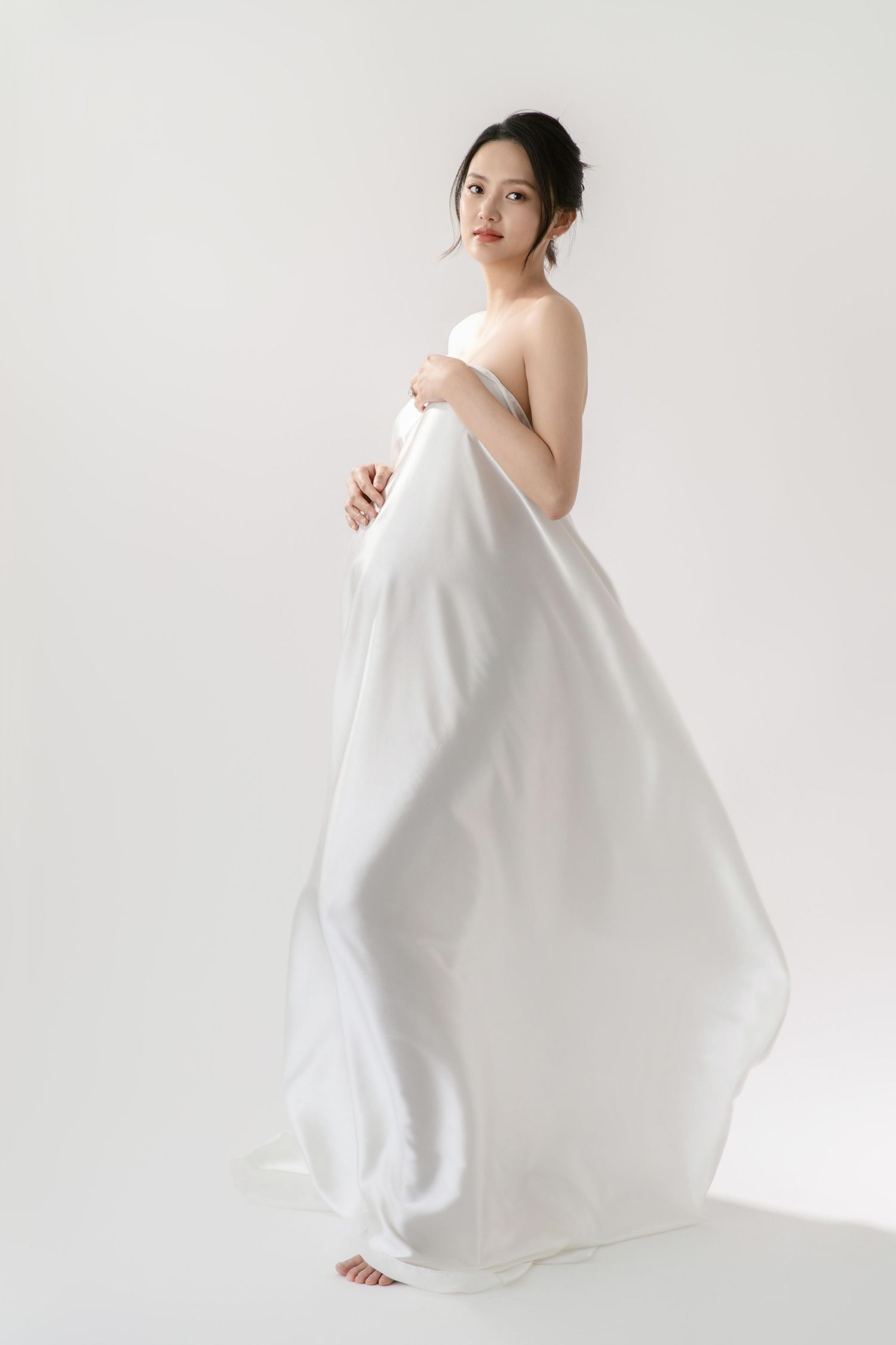 Graceful maternity portrait with billowing white fabric against white backdrop.