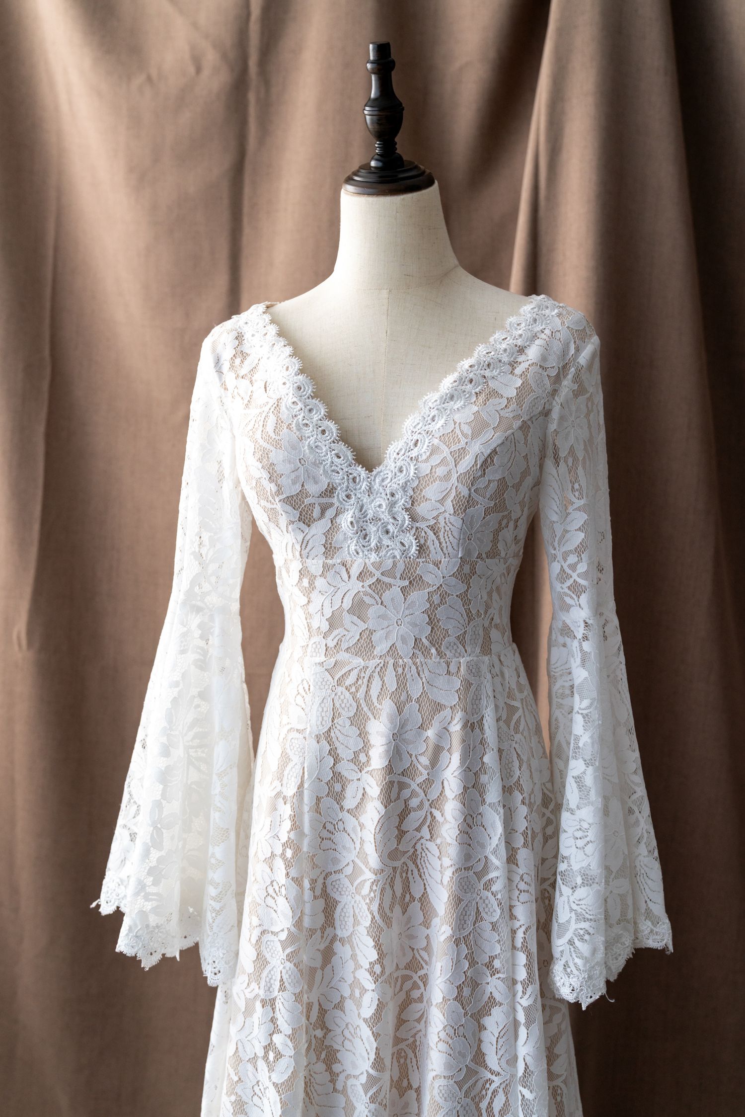 White lace wedding dress with bell sleeves displayed on mannequin against brown backdrop.