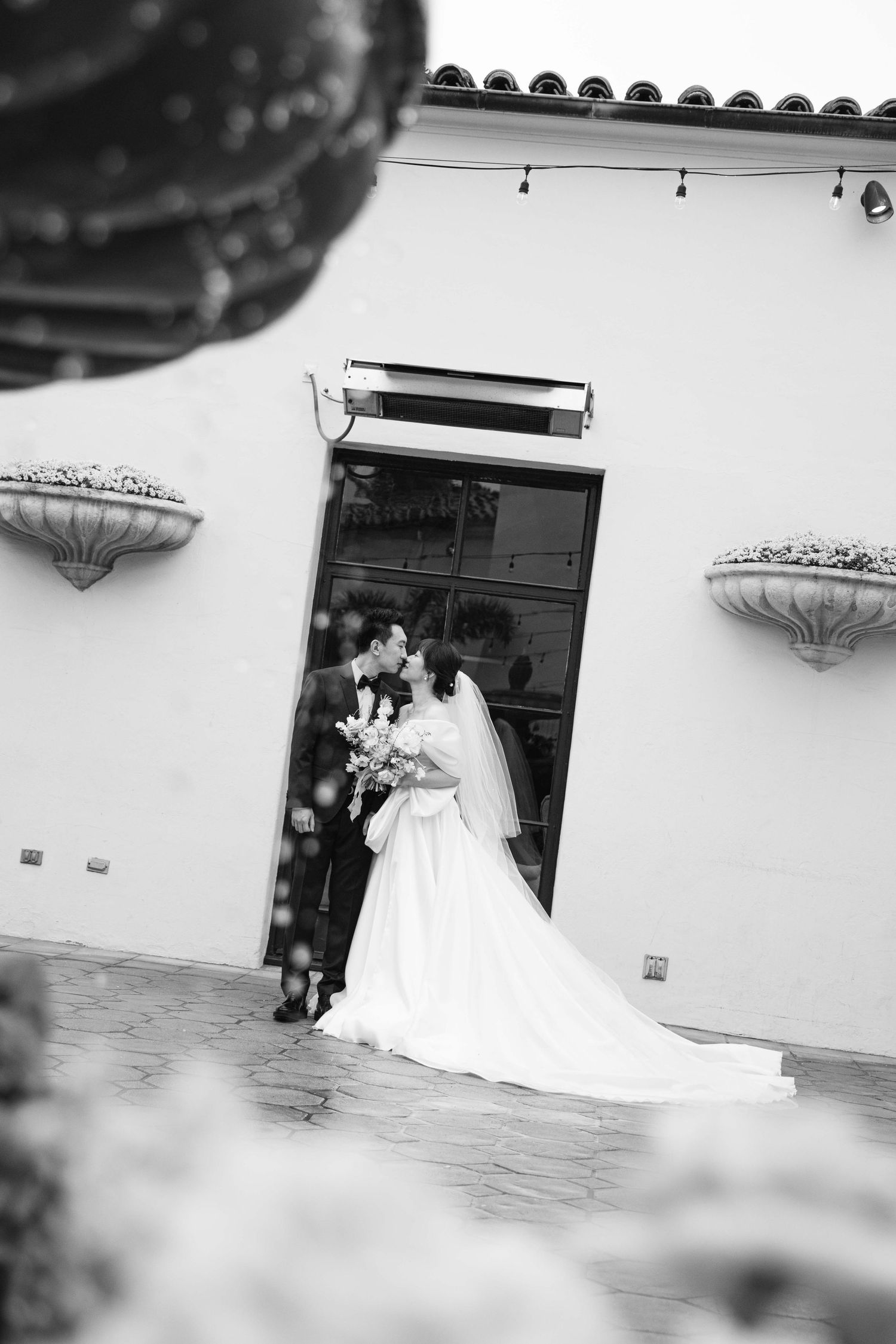 A couple shares a romantic moment in their wedding attire against a white Spanish-style building exterior.
