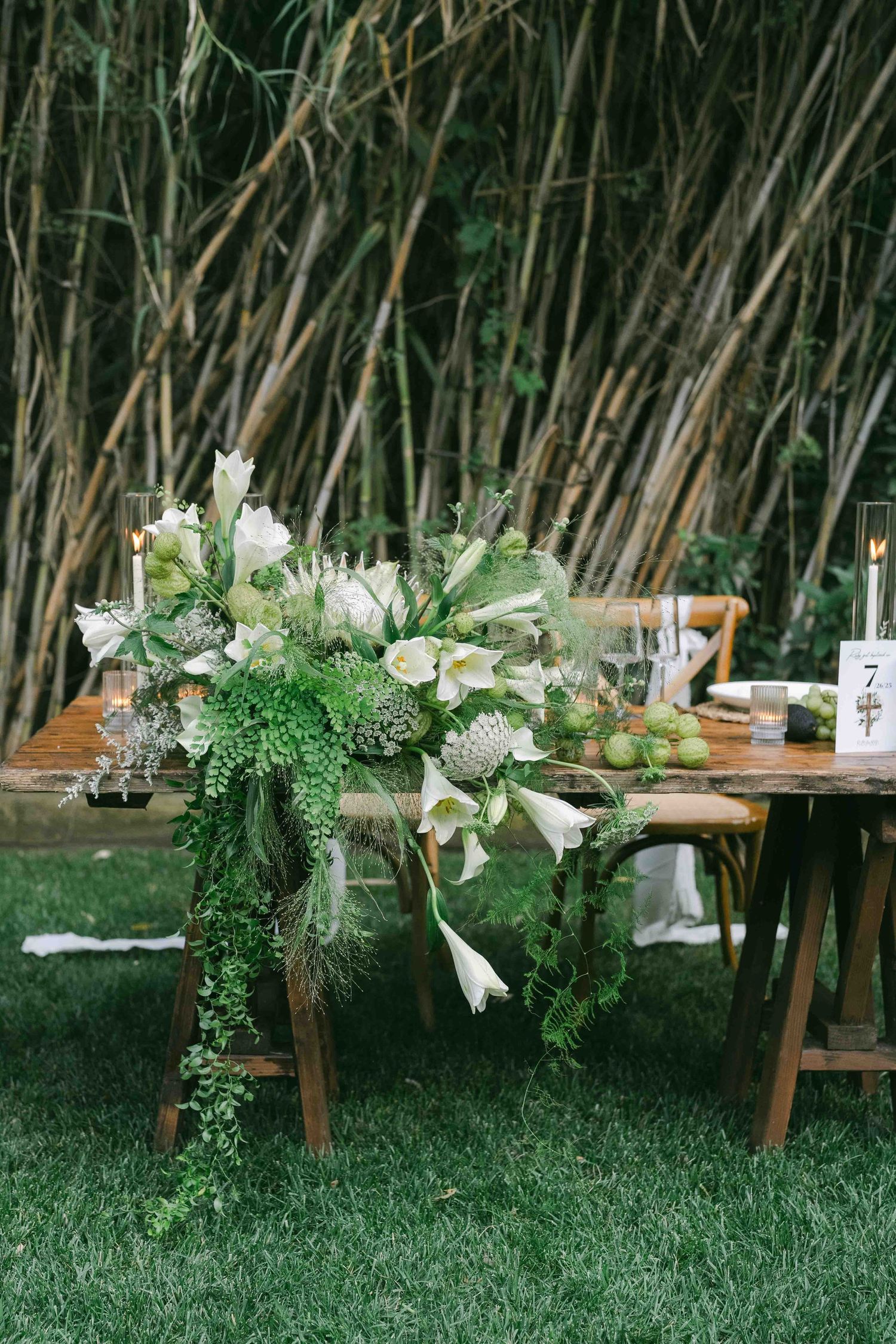 Lush green and white floral arrangements adorning wooden dining tables against tall grass backdrop.