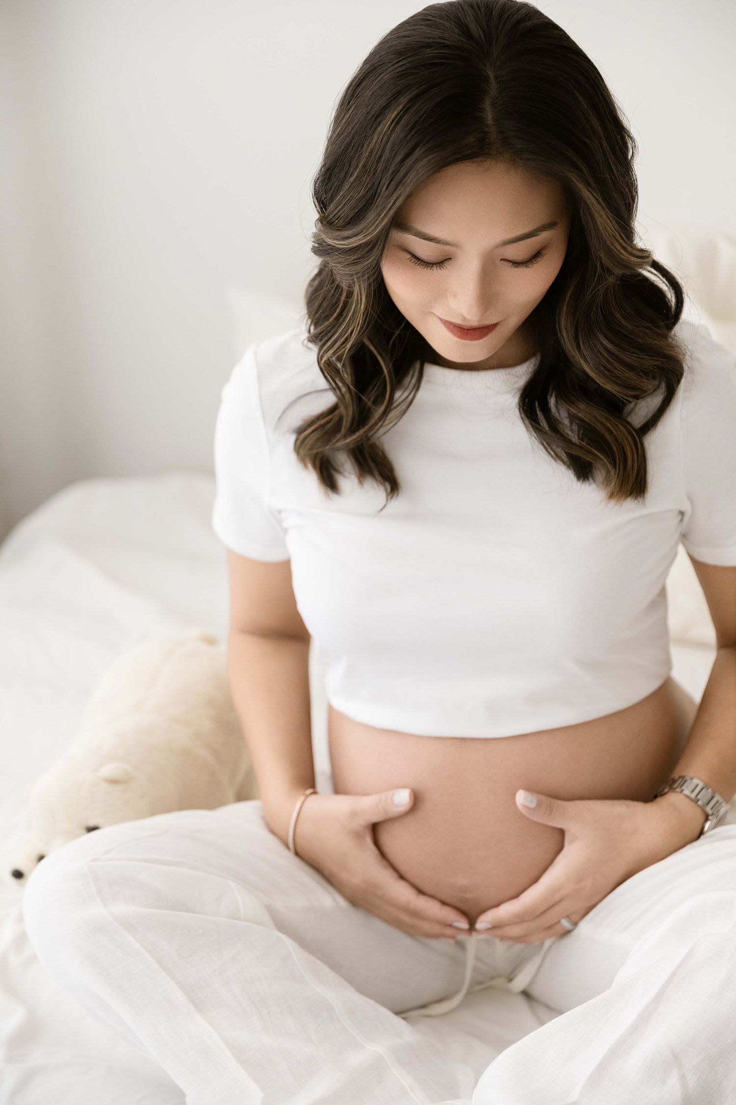 Expectant mother in white clothing gently touching her baby bump on white bedding.