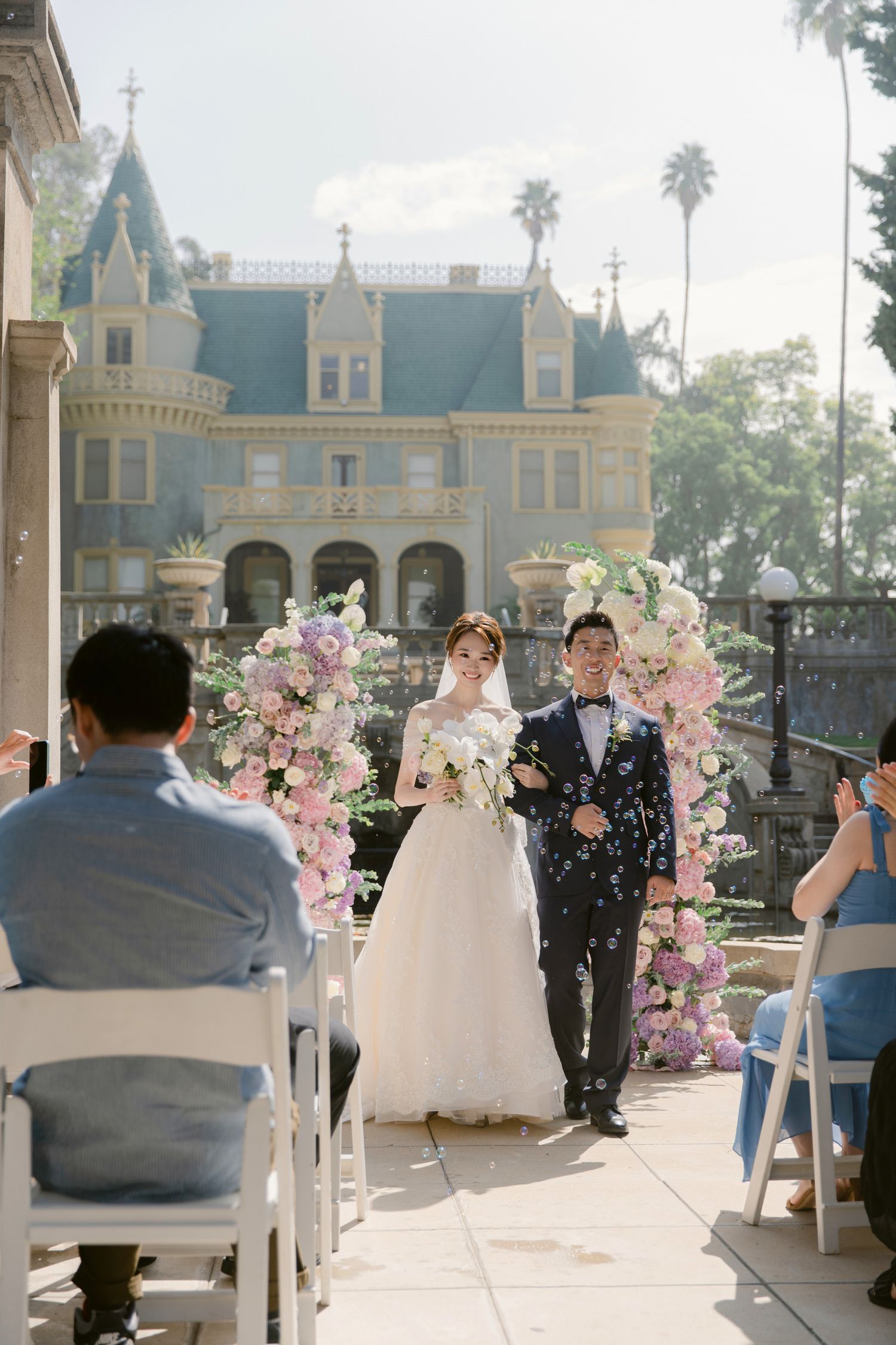 Wedding ceremony with guests seated in white chairs and pink floral decorations.